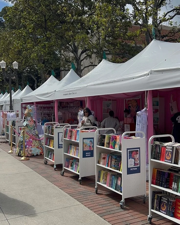 Outdoor book sale with white tents and bookshelves labeled 'Beach Reads' and 'Fiction.' People browsing, colorful decorations, trees and lamp posts in the background.