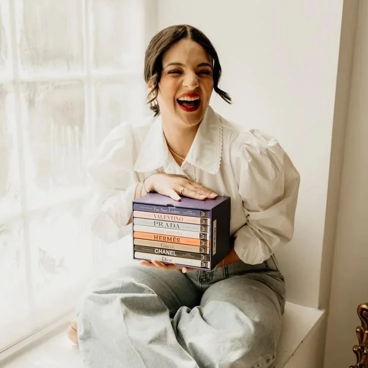 A woman sitting on a windowsill, smiling and holding a stack of books about luxury fashion brands.
