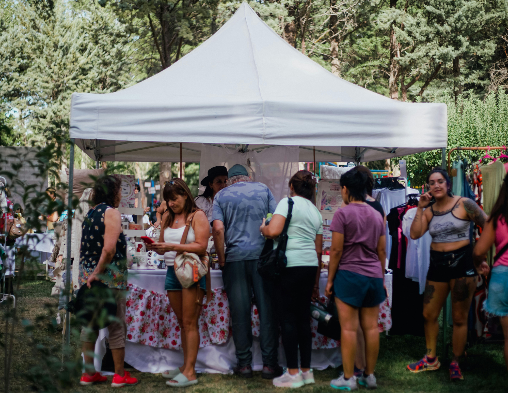 People gathered around a booth under a white canopy tent at an outdoor market or festival, with trees and greenery in the background