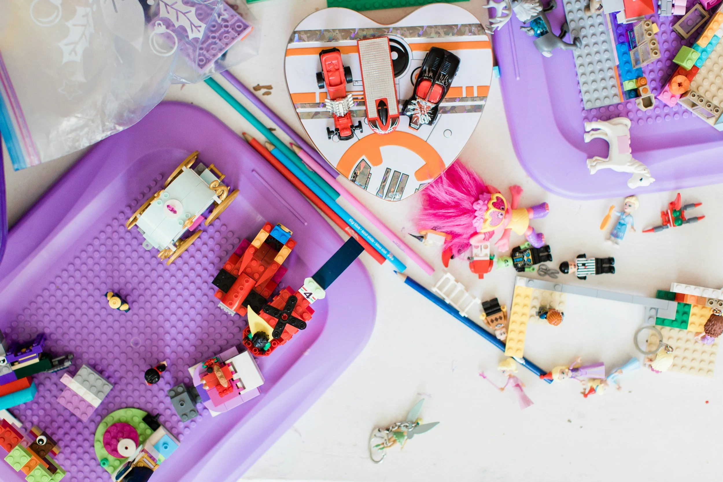 A messy table with Lego and toy figures, purple storage bins, scattered construction pieces, a reflective heart-shaped mirror, and various small toys including a doll with pink hair and a rainbow-colored skirt.