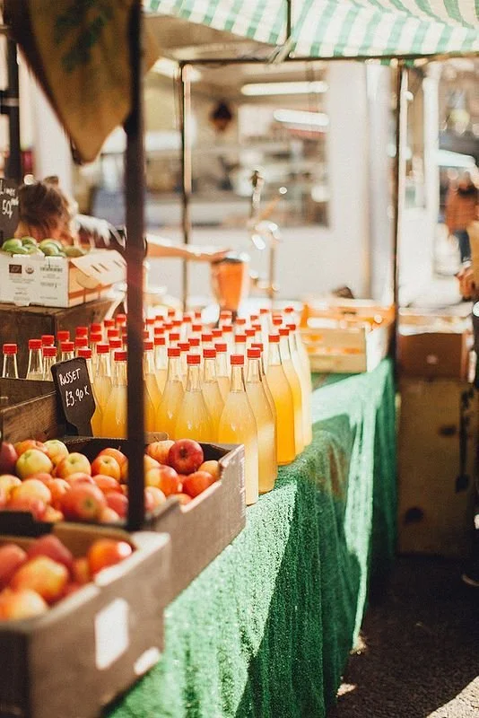 A fruit stand at an outdoor market with apples in a wooden crate and bottles of orange juice on a green tablecloth.