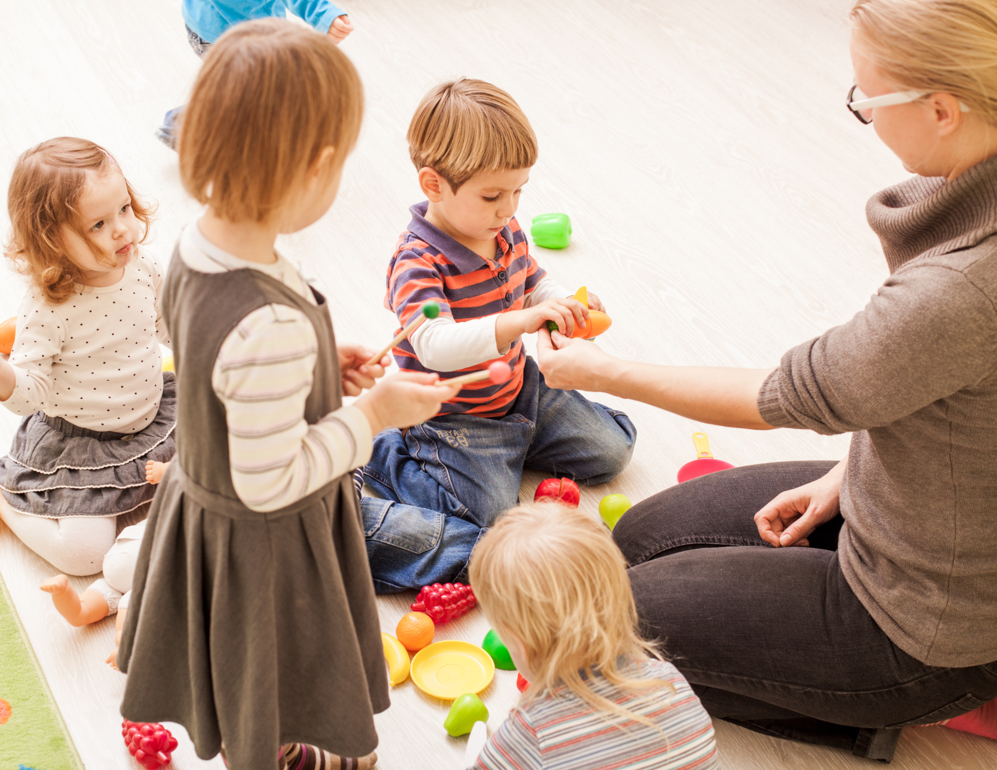 Children playing with colorful plastic toy food and kitchen utensils while a woman watches. The children sit on a light wood floor surrounded by toy fruits and vegetables.
