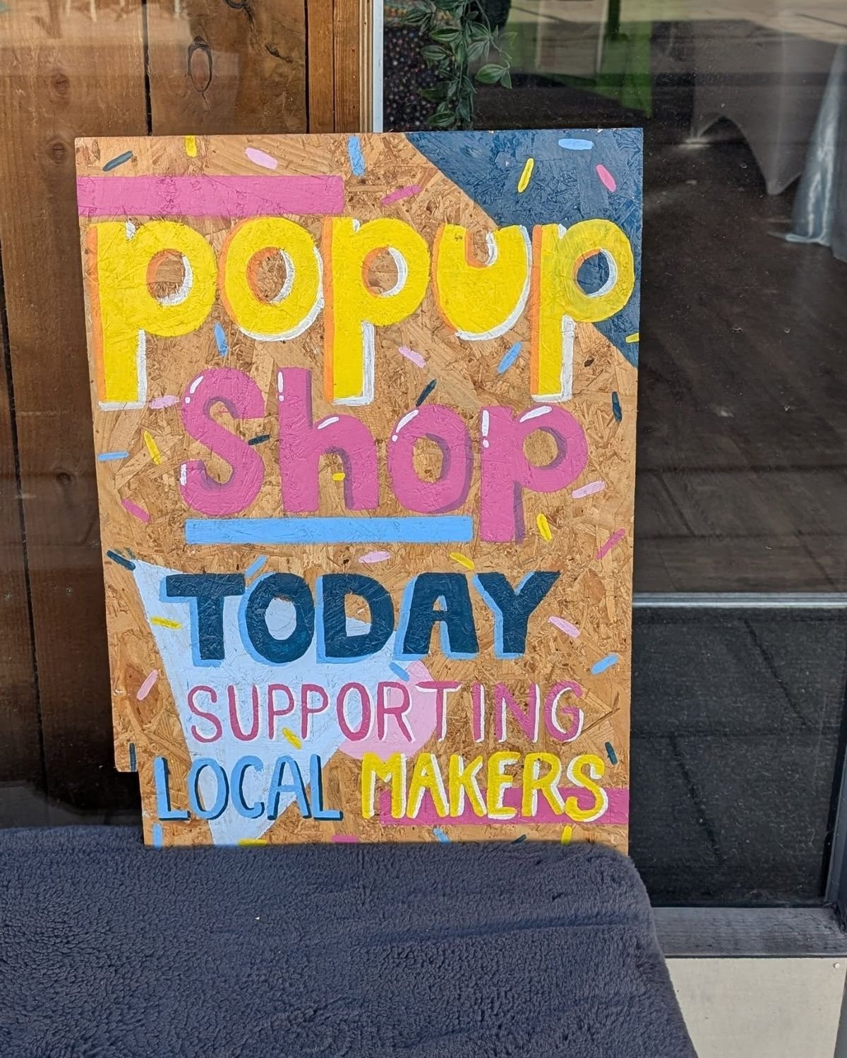 Colorful handmade sign on a wooden board promoting a pop-up shop supporting local makers, with bright pink, yellow, blue, and white lettering and confetti decorations.