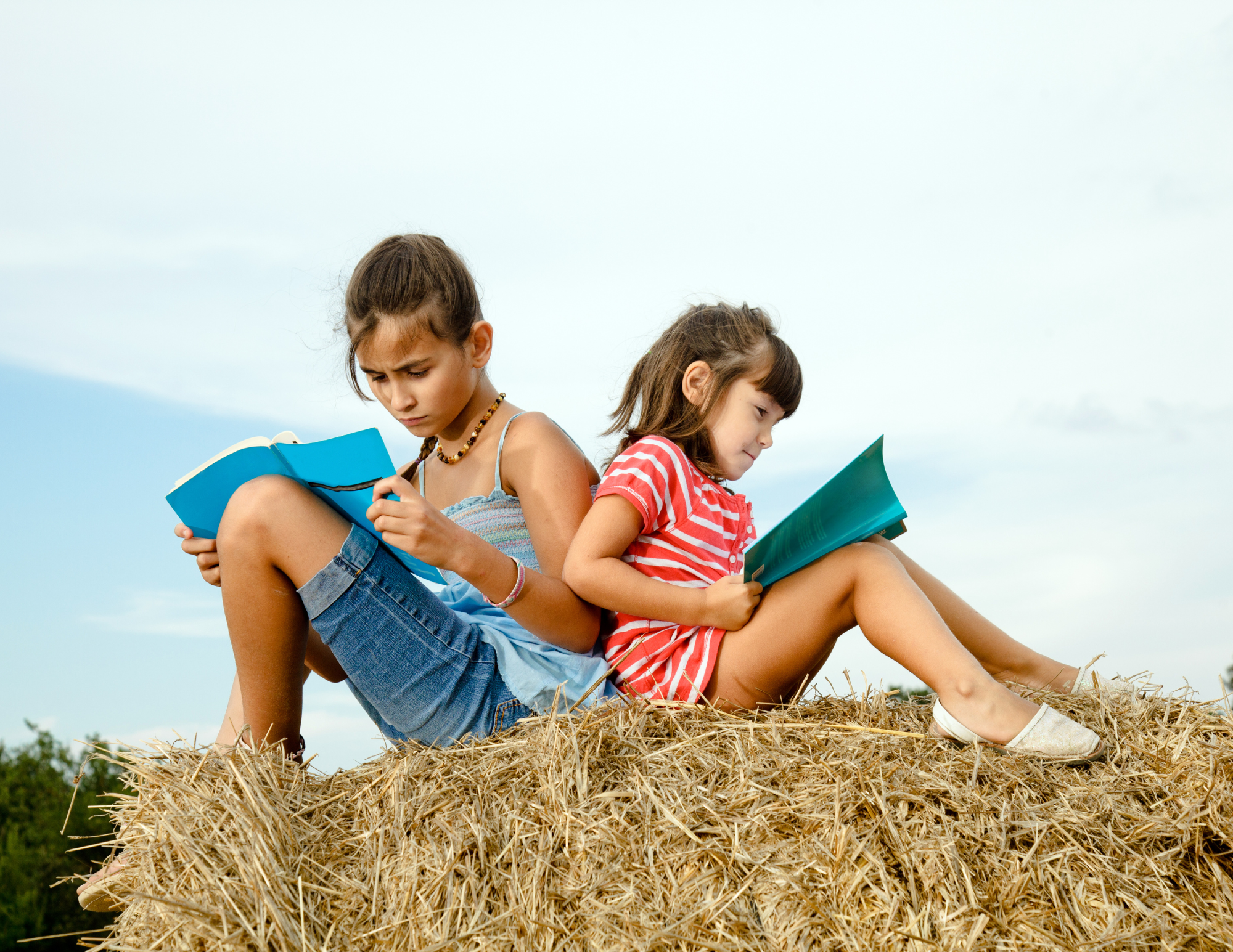 Two young girls sitting on a hay bale outdoors, reading books, with a clear sky background.