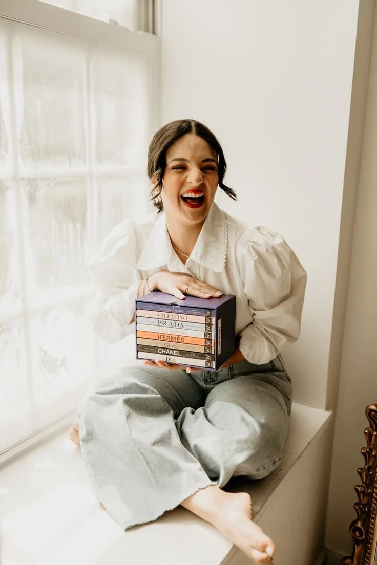 A woman with short dark hair and red lipstick sitting on a windowsill, smiling and holding a stack of books about luxury brands.