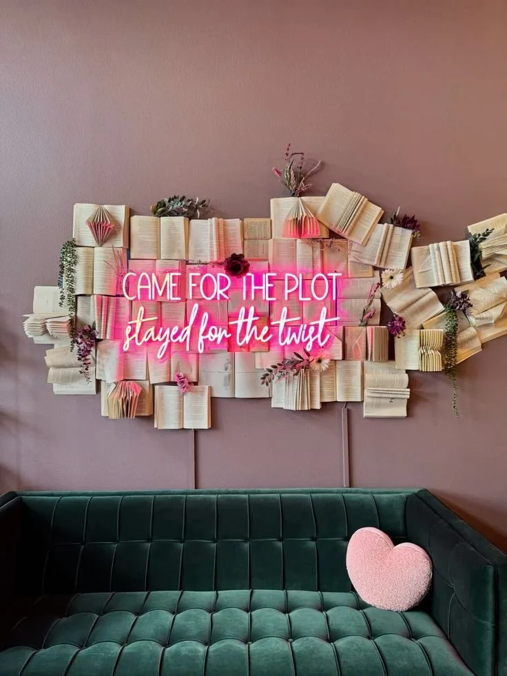Decorative wall made of open books with a pink neon sign that reads 'CAME FOR THE PLOT stayed for the twist,' surrounded by flowers and greenery, with a dark green sofa and a pink heart-shaped pillow in front.