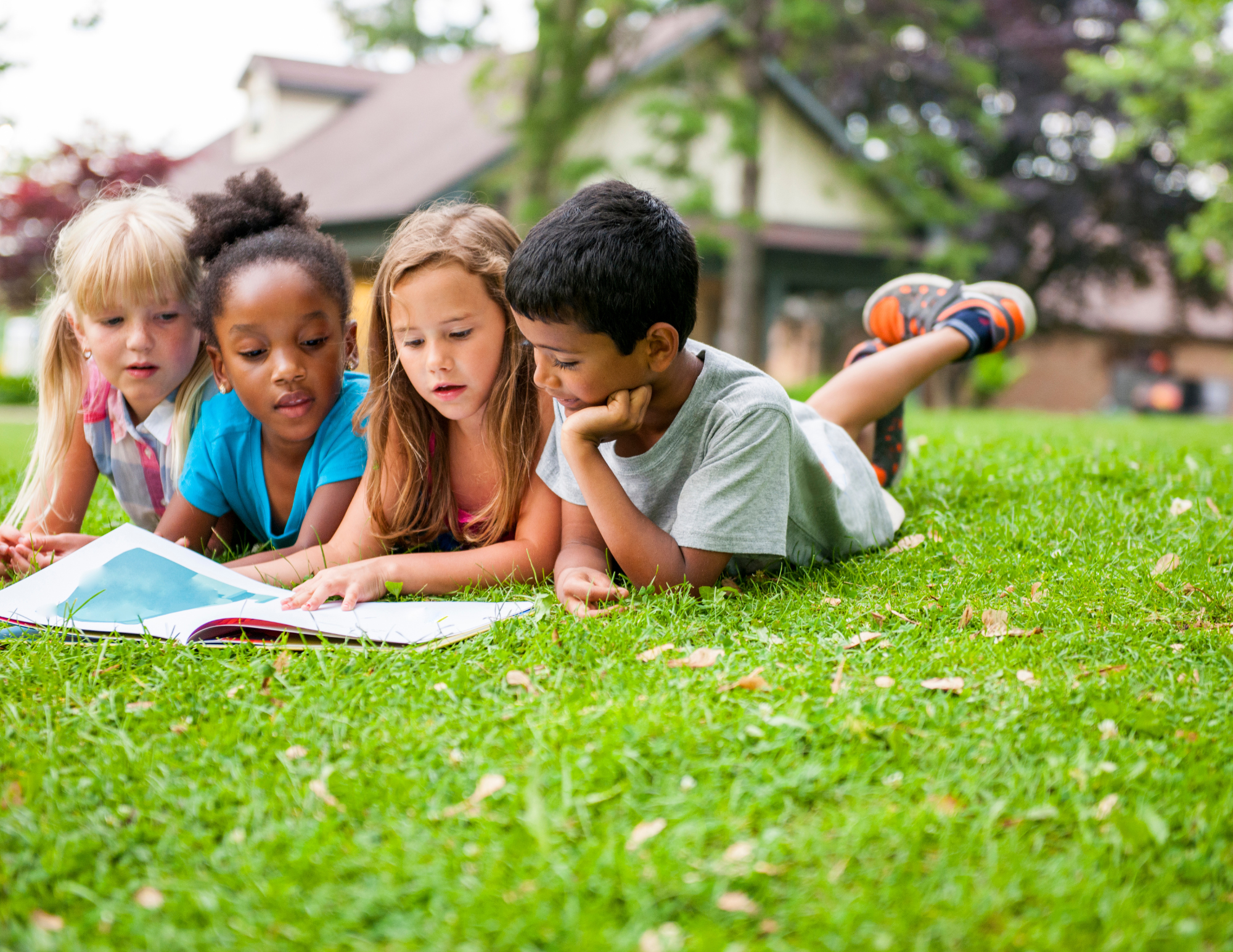 Four children, two girls and two boys, lying on the grass and looking at a book together in a park with trees and houses in the background.
