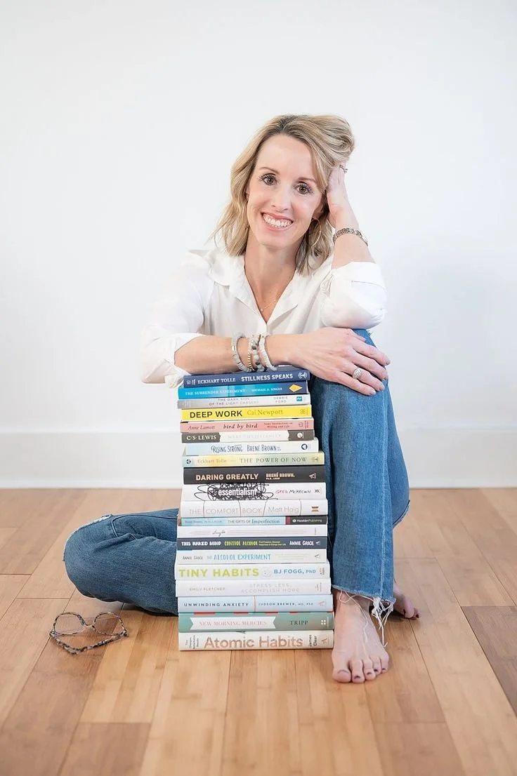 A woman with blonde hair sitting on the floor with crossed legs, smiling, and leaning on a stack of books with titles related to mindfulness, self-improvement, and habits. She is wearing a white blouse, jeans, and has glasses on the floor beside her.