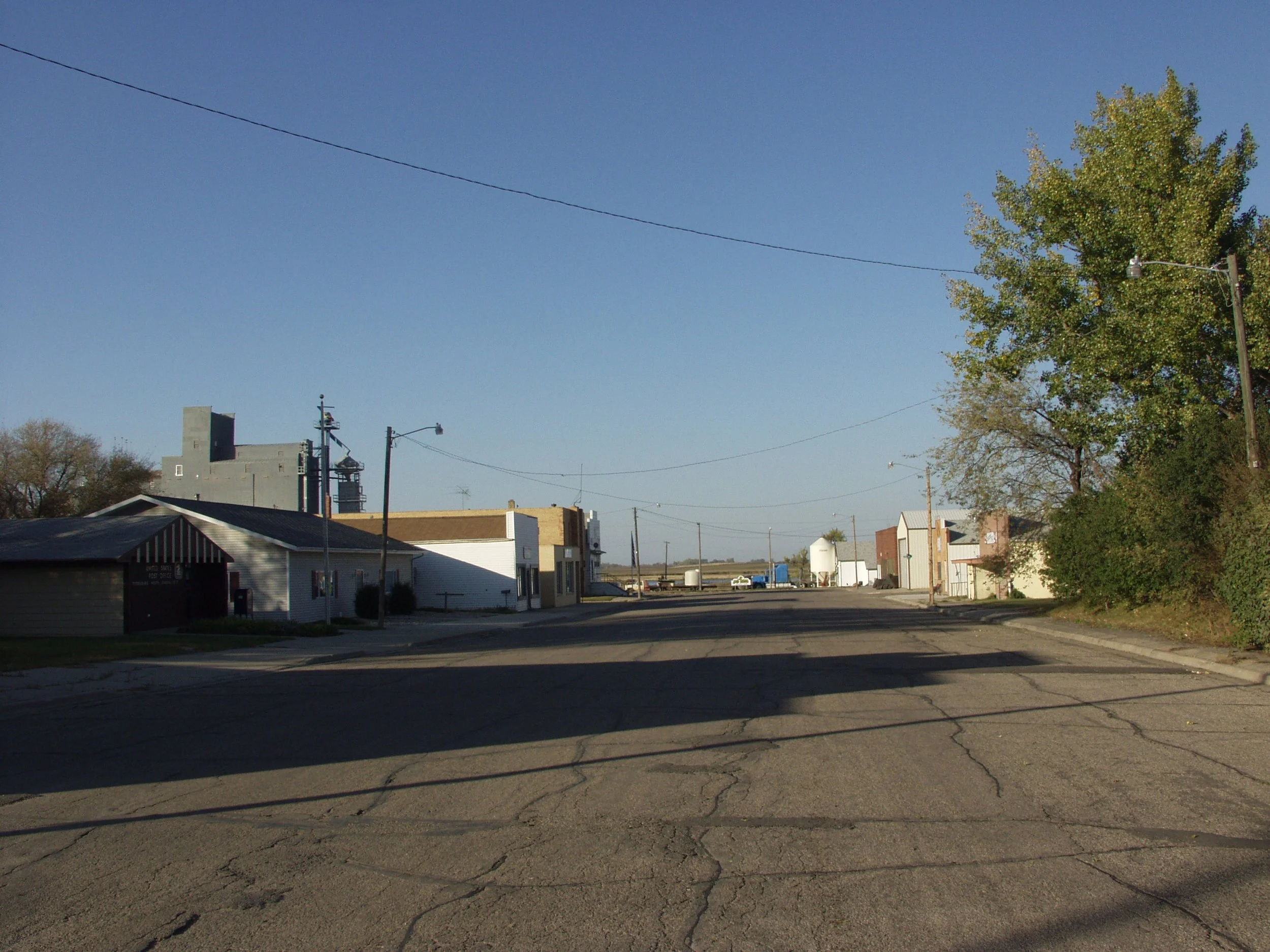 A small town street with buildings on both sides, utility poles, a tree with green leaves, and a clear blue sky.