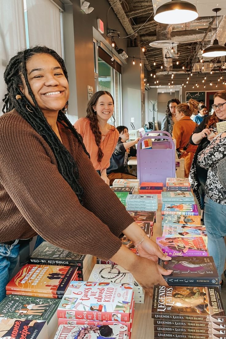 People browsing and shopping at a book sale or bookstore with tables full of books, in a cozy, industrial-style space with hanging lights and large windows.