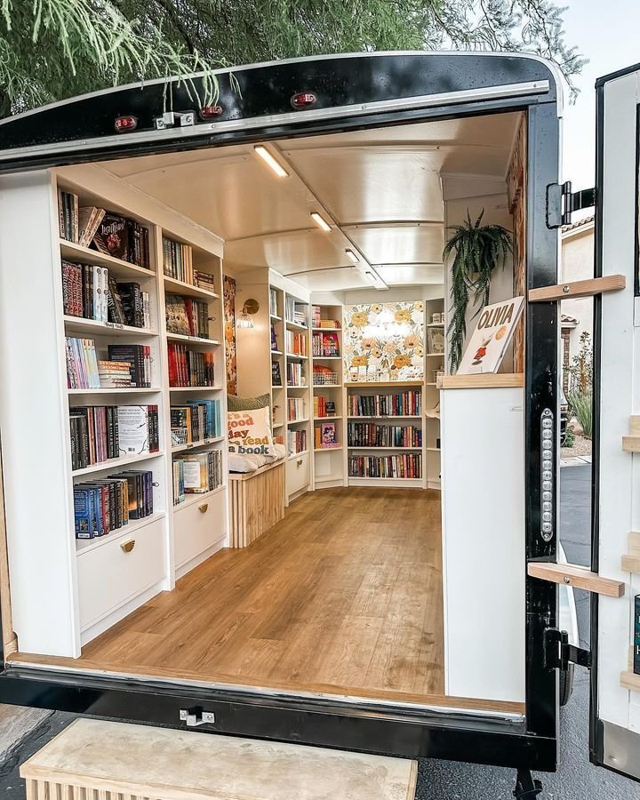 Inside a black mobile library or book lending trailer with white shelves filled with books, a wooden floor, and a cozy nook with cushions and decorative pillows, including one that says 'Read a Book'.
