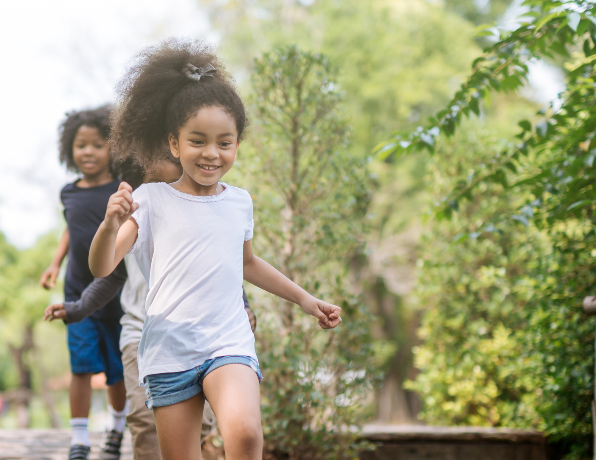 Three young girls are running outdoors on a sunny day, surrounded by green trees and bushes. The girl in front has curly hair tied back with a black ribbon, wearing a white t-shirt and denim shorts, smiling happily. The other two girls are behind her, also smiling and running along the same trail.