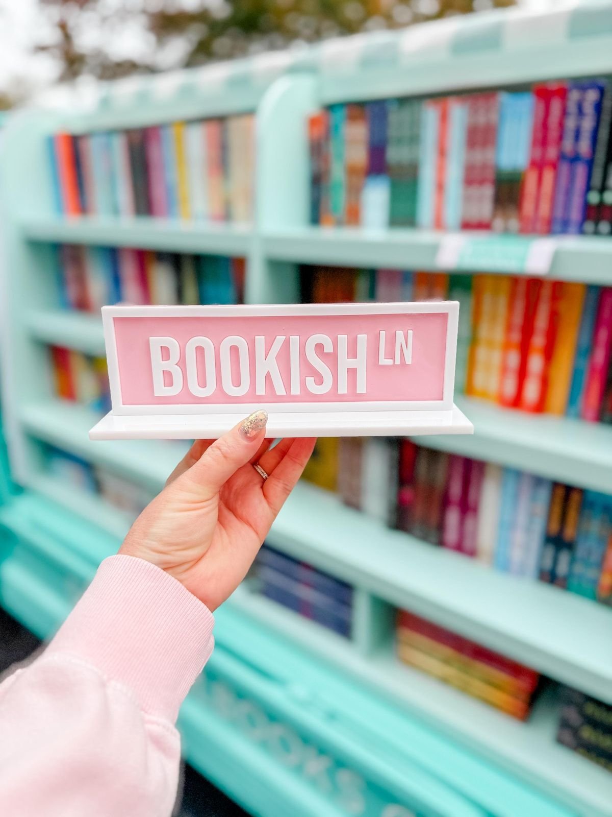 A person holding a sign that says 'BOOKISH LN' in front of a bookshelf filled with colorful books.