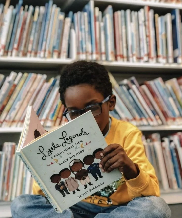 A young boy with glasses, wearing a yellow sweatshirt, sitting in a library and reading a book titled 'Little Legends: Extraordinary Men in Black History' by Vashti Harrison.