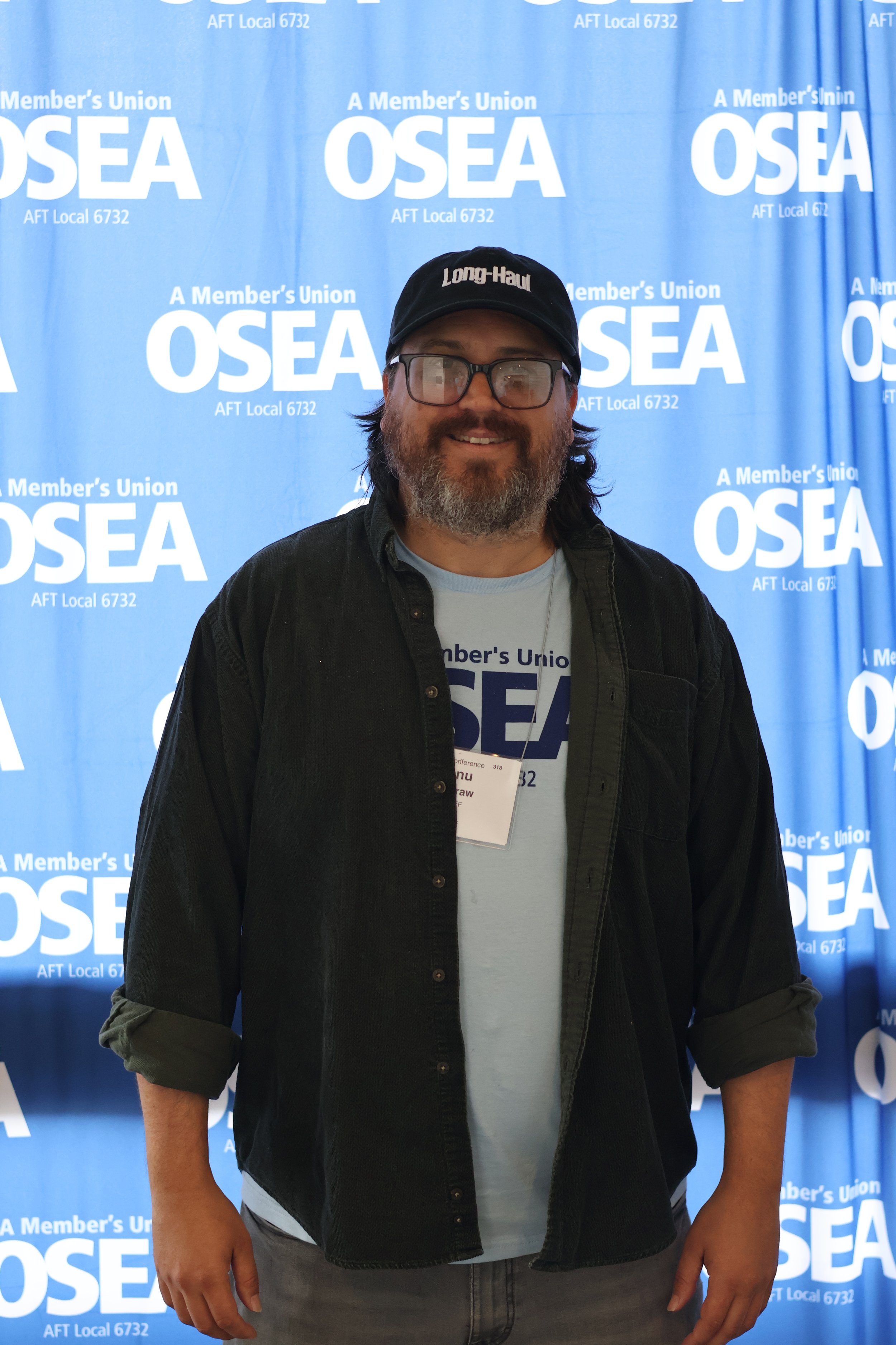A man with glasses and a beard smiling in front of a blue backdrop with white text that reads 'A Member's Union OSEA' and 'AFT Local 6732'. He is wearing a black cap with white text, a black shirt over a white T-shirt, and gray pants.