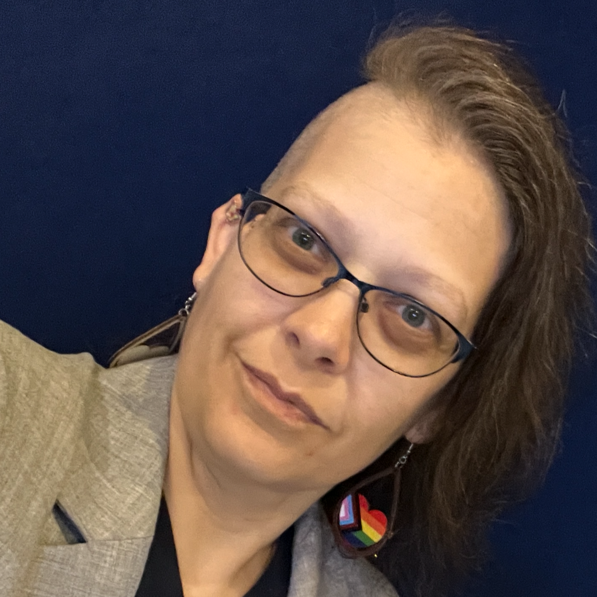 A woman with short, styled hair wearing glasses and rainbow pride earrings, smiling against a dark blue background.