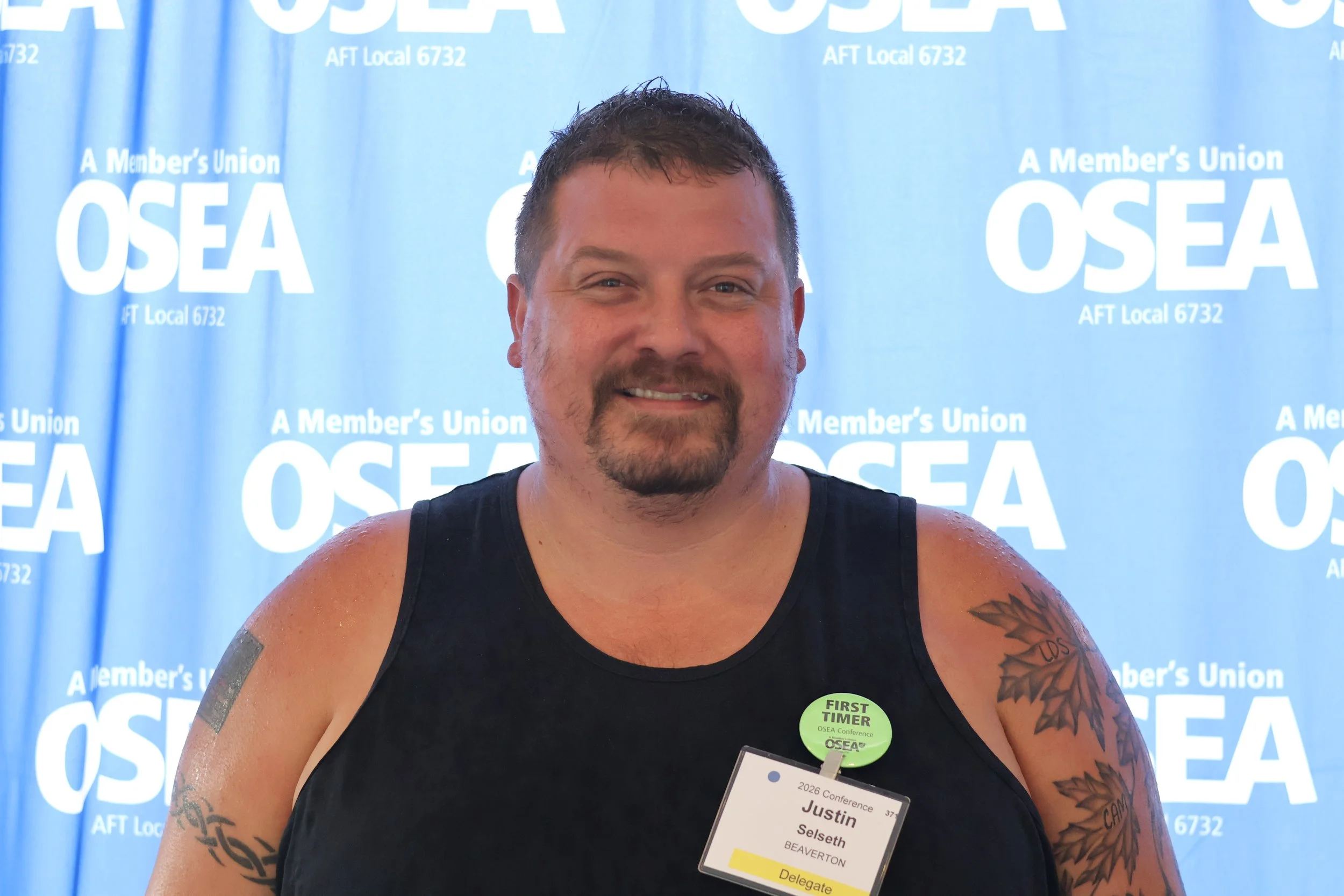 A man with short dark hair, a goatee, and tattoos on his arms, smiling and wearing a black tank top, standing in front of a blue backdrop with white text that reads 'A Member's Union OSEA' and other related phrases.
