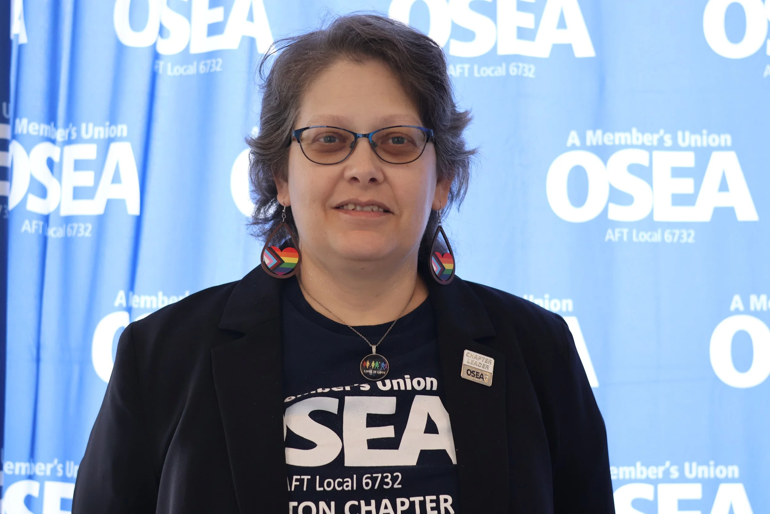A woman with short, styled hair wearing glasses and rainbow pride earrings, smiling against a dark blue background.