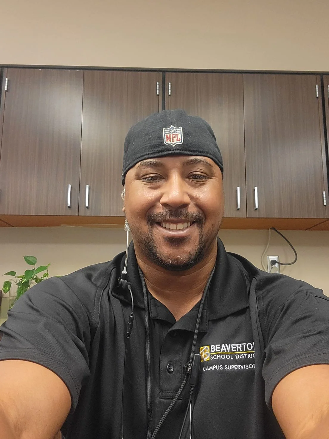 A man smiling for a selfie, wearing a beanie with NFL logo, a black shirt with Beaver County School District logo, and a headset around his neck, in an indoor setting with cabinets and a plant in the background.