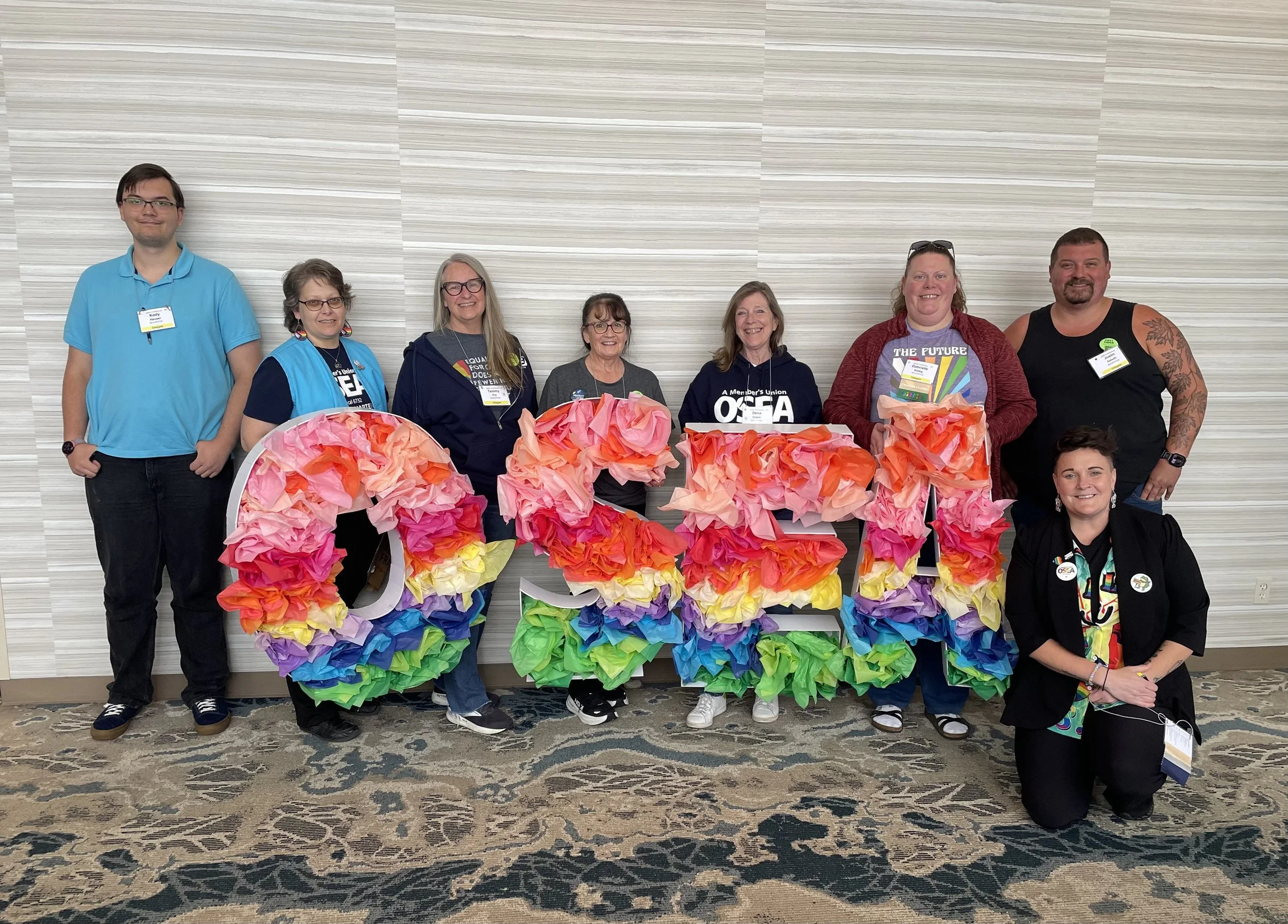A group of seven diverse people standing behind a rainbow-colored paper flower display against a light gray striped wall. They are smiling and wearing casual clothing with some wearing badges or pins. The scene appears to be at a conference or event.
