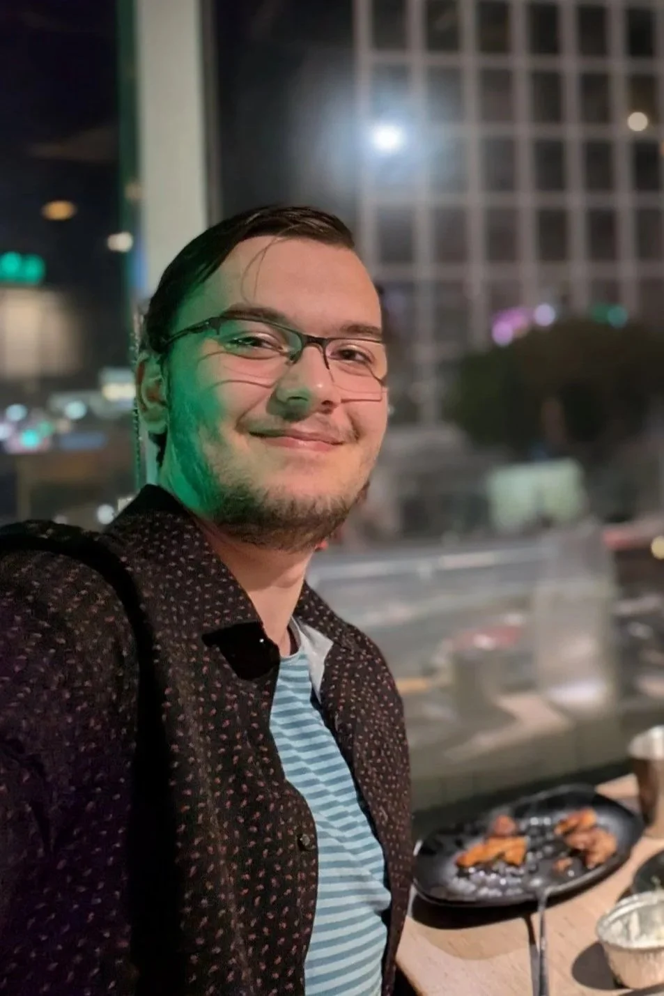 A smiling man with glasses and a goatee, wearing a dark patterned shirt and a striped t-shirt, stands outdoors at night near a grill with food on it, with city buildings and lights in the background.