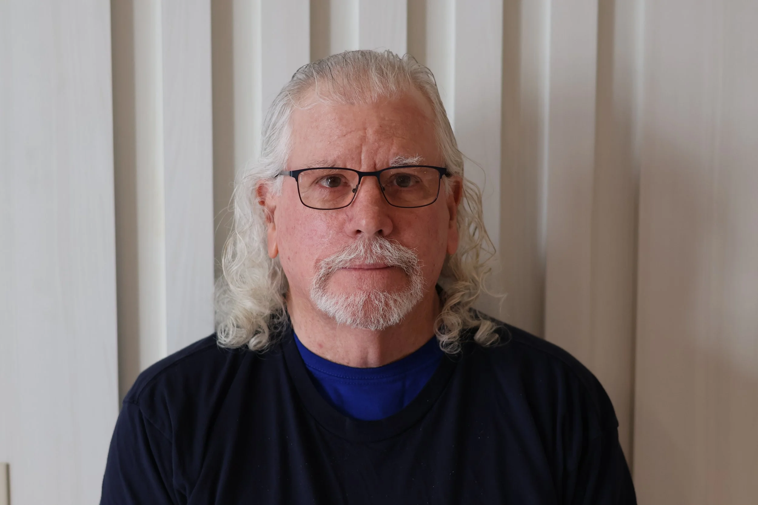 Close-up of a man with long, curly white hair and a beard, wearing glasses, standing in front of vertical white blinds.
