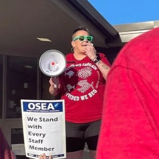 A woman protesting outdoors, holding a megaphone and a sign that says 'OSEA We Stand with Every Staff Member.' She is wearing sunglasses, a red shirt, and has a tattoo on her arm.