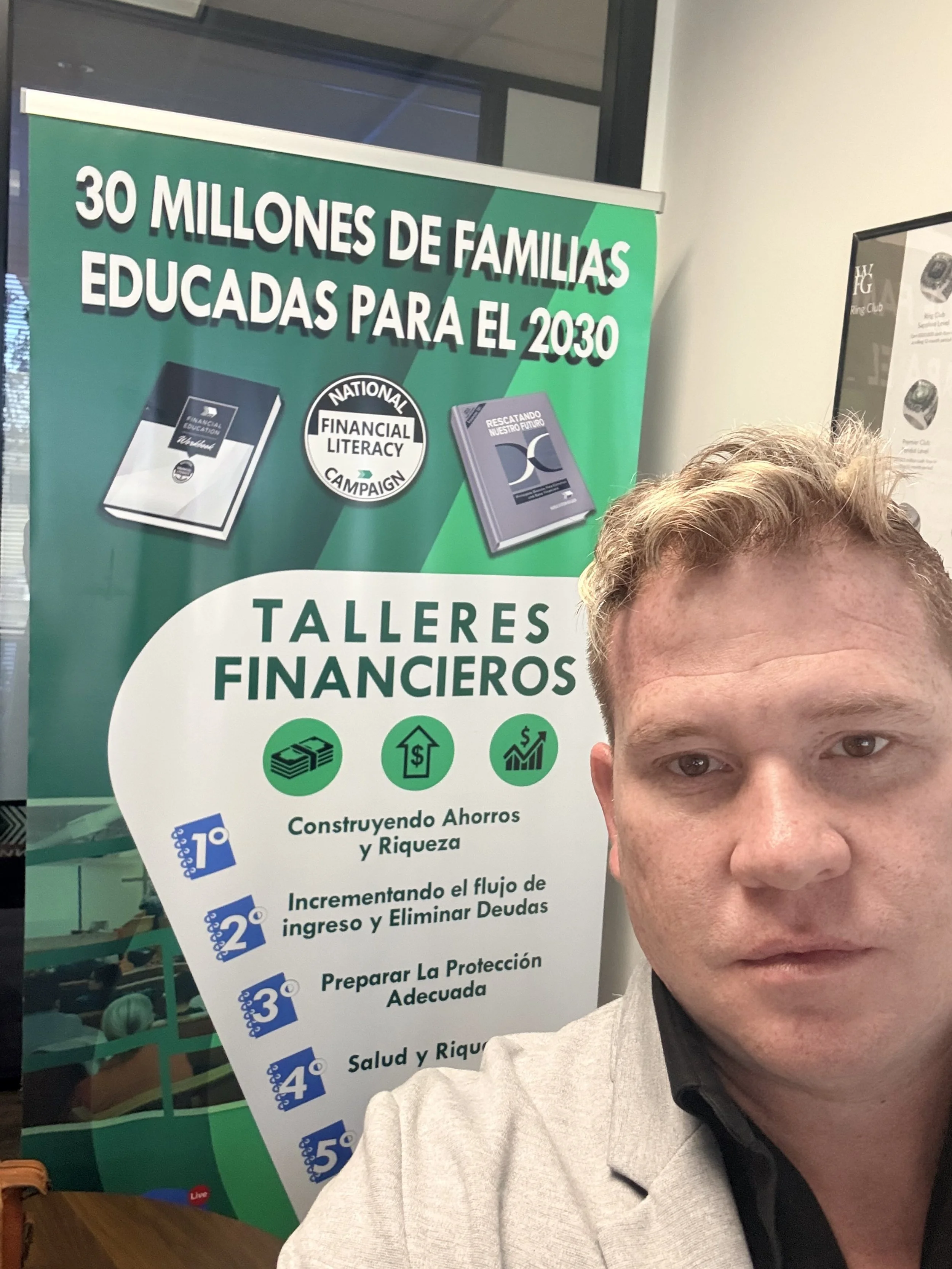 Hombre con chaqueta gris y camisa negra frente a un cartel en un evento o conferencia sobre talleres financieros y alfabetización financiera.