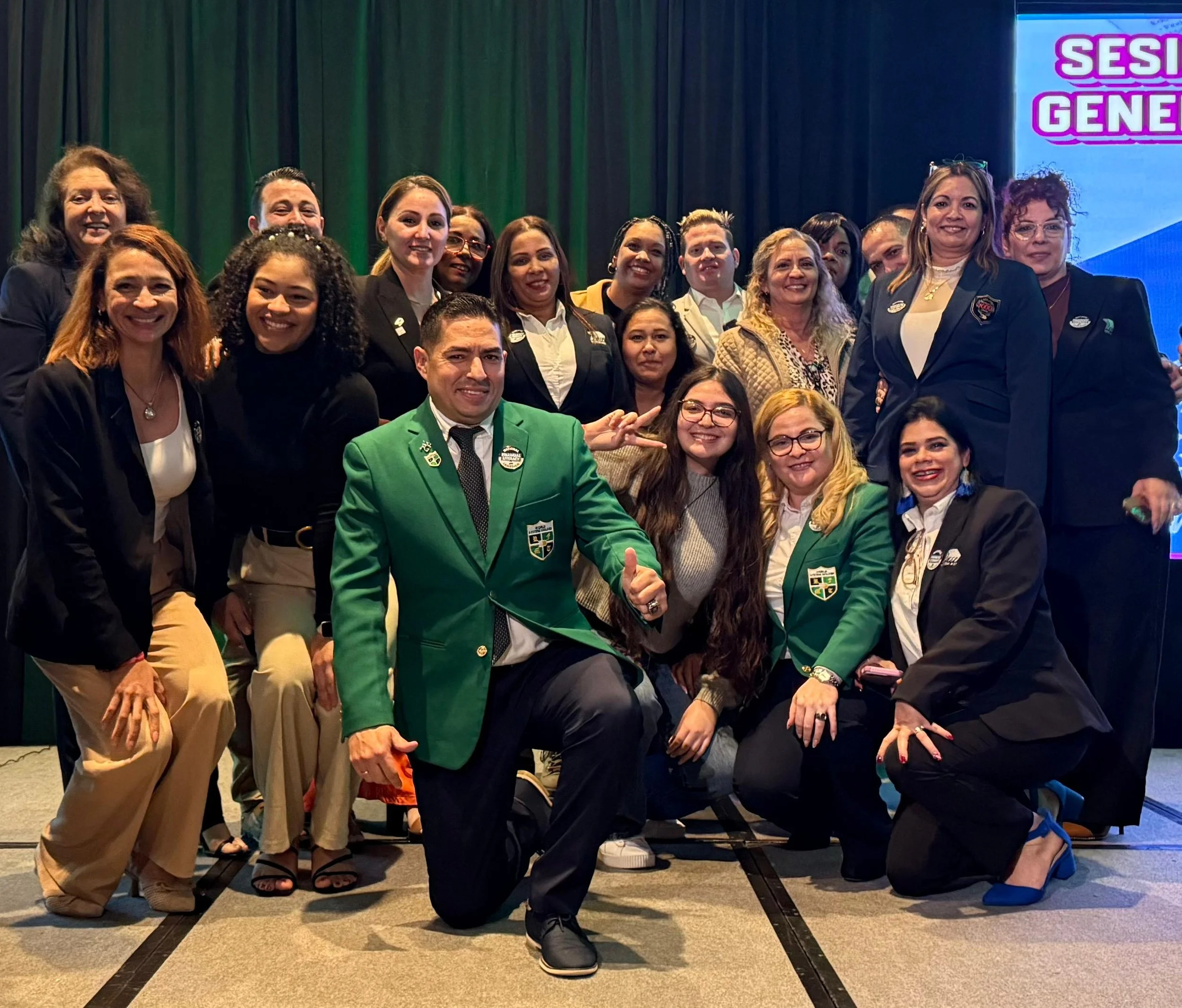 Grupo de personas en un evento, con algunos usando trajes formales y otros en blazer con insignias, en un escenario con pantalla que dice 'SESI GENE' en letras moradas y rosa.