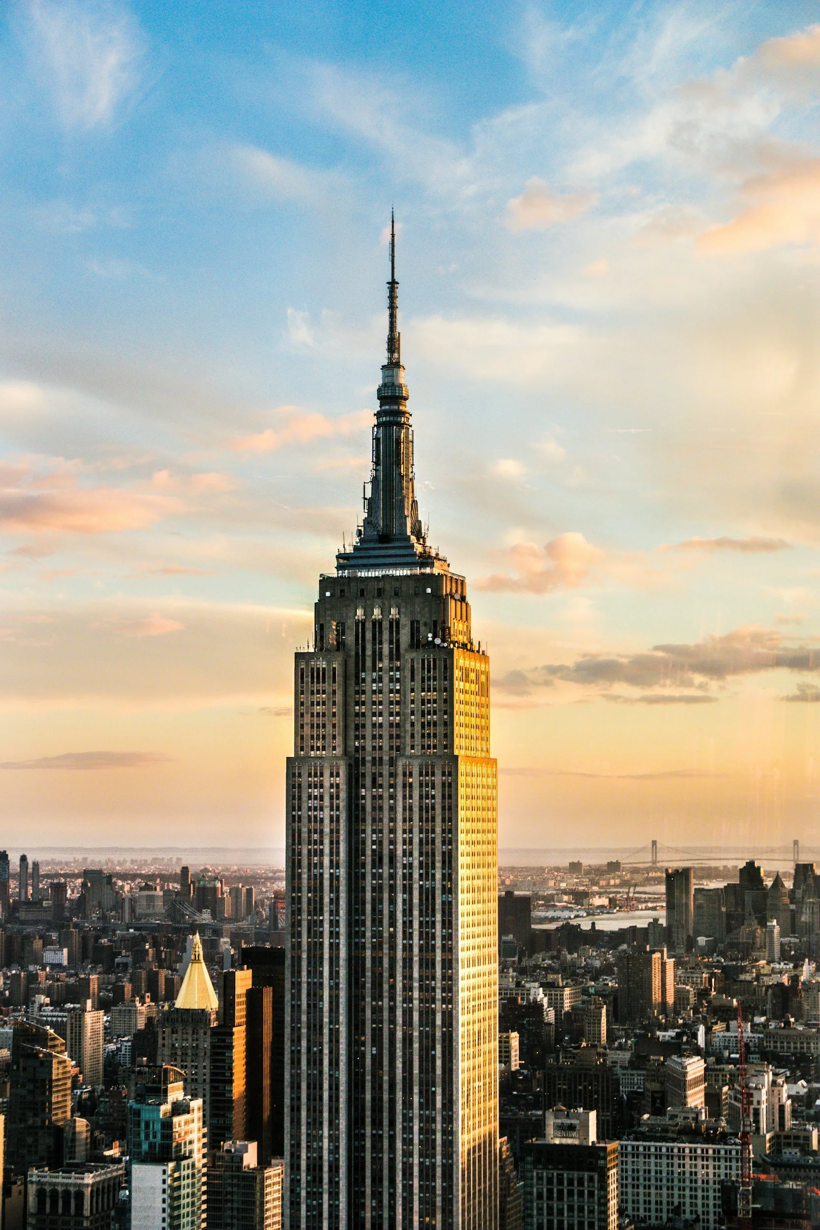 The Empire State Building towering over New York City with a partly cloudy sky and sunset lighting.
