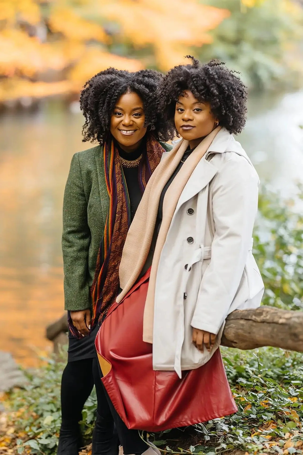 Two women with curly black hair standing outdoors near water surrounded by fall foliage, smiling and dressed in layered coats and scarves.