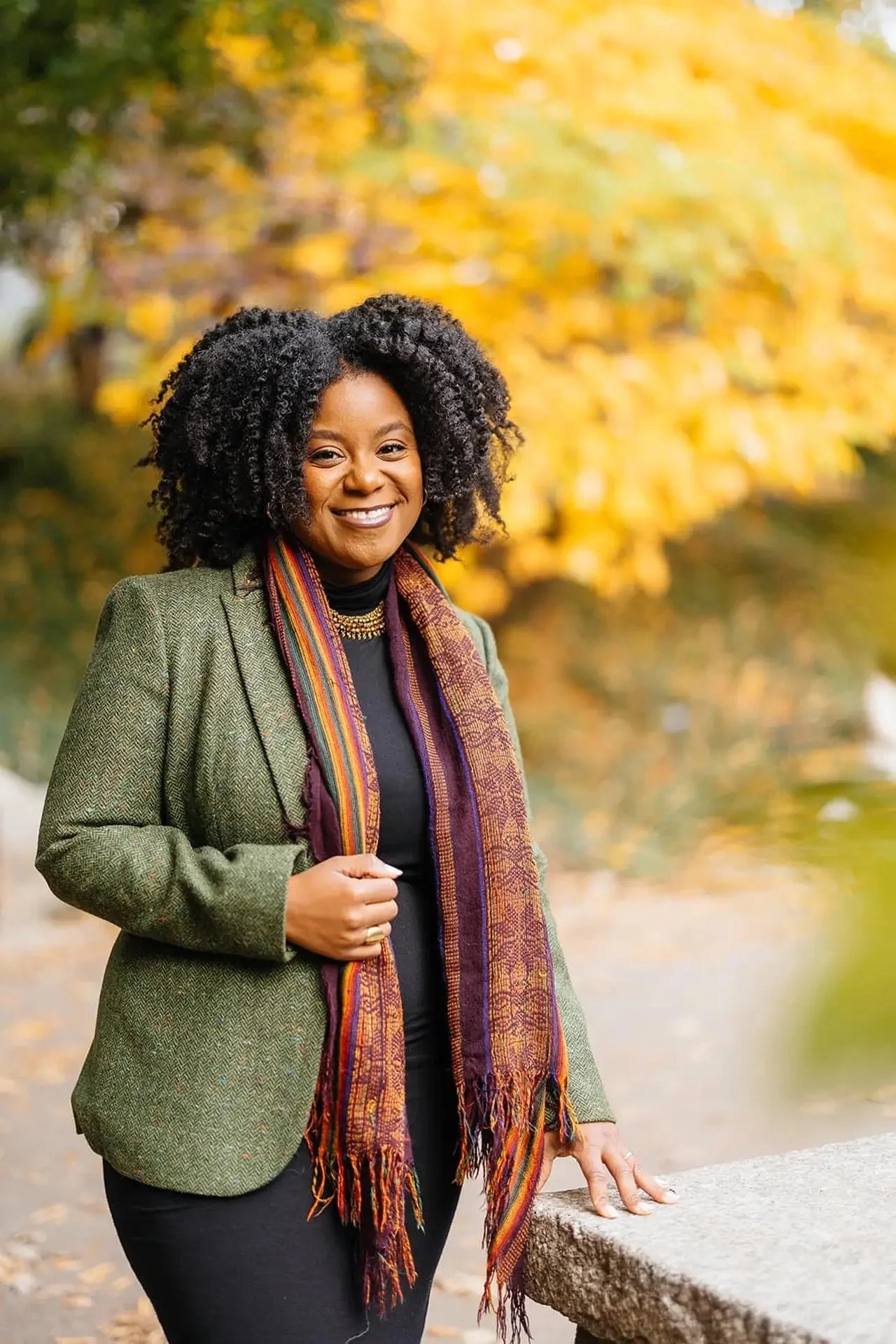 A woman with curly black hair smiles outdoors during autumn, wearing a green blazer, black top, and a multicolored scarf, standing near a stone railing with fall foliage in the background.