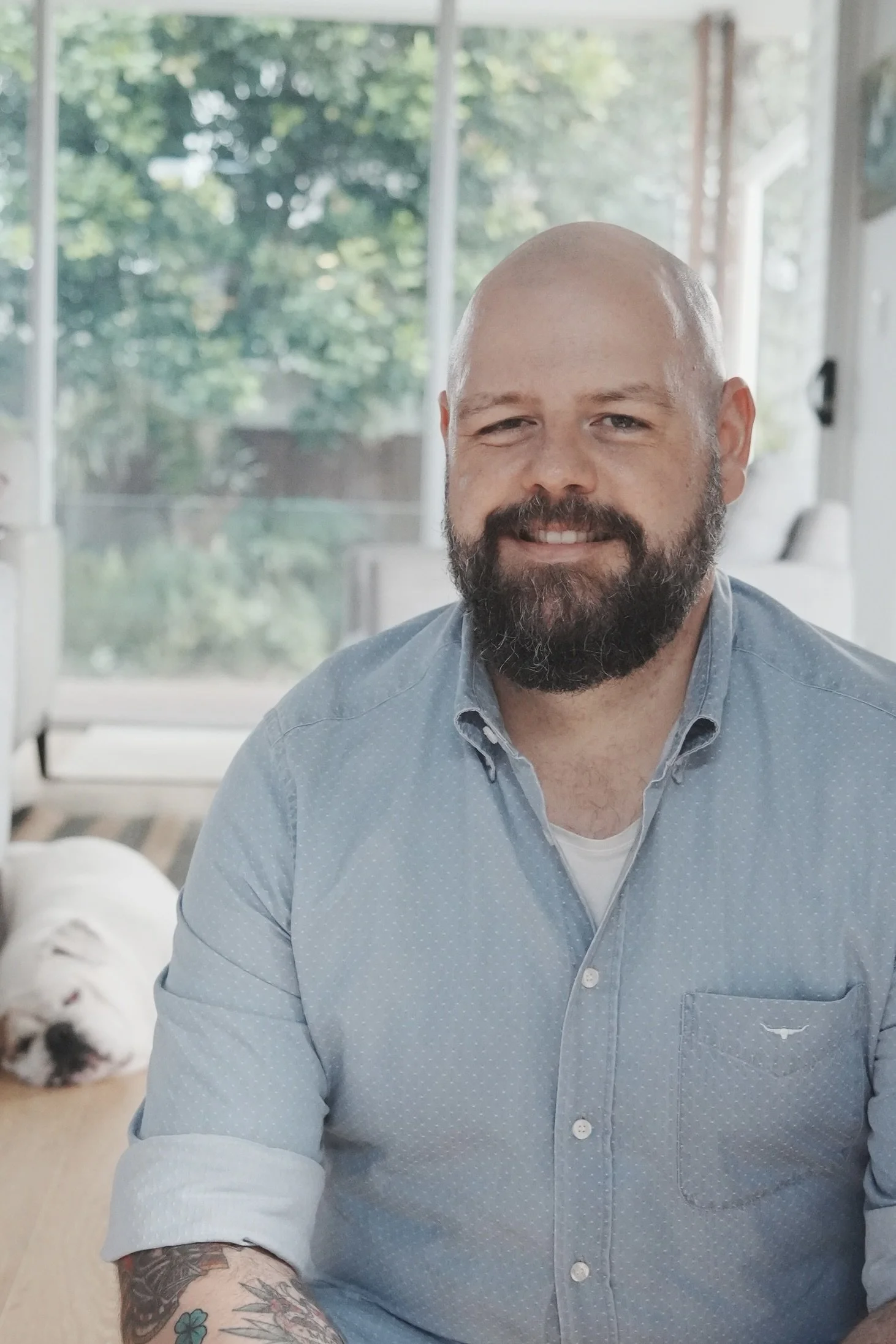 A smiling man with a beard and tattoos on his arm, wearing a light blue button-up shirt, sitting indoors with a dog sleeping on the floor in the background.