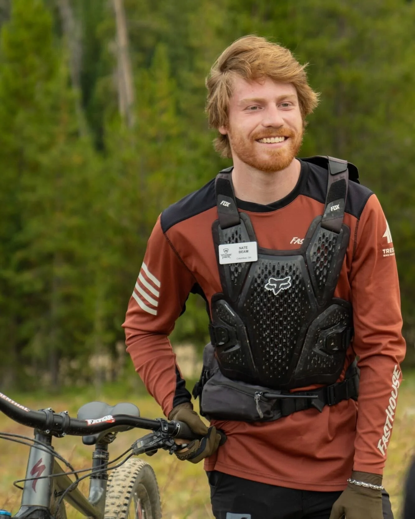 Man in mountain biking gear standing next to a bicycle outdoors in a forested area.