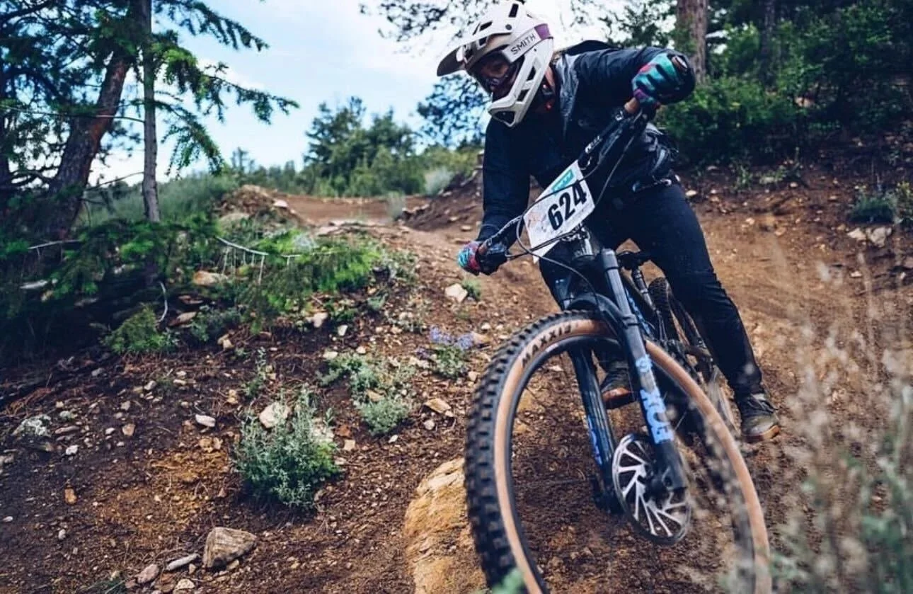 A mountain biker wearing a full-face helmet, goggles, and black gear navigates a dirt trail on a slope surrounded by trees and greenery.