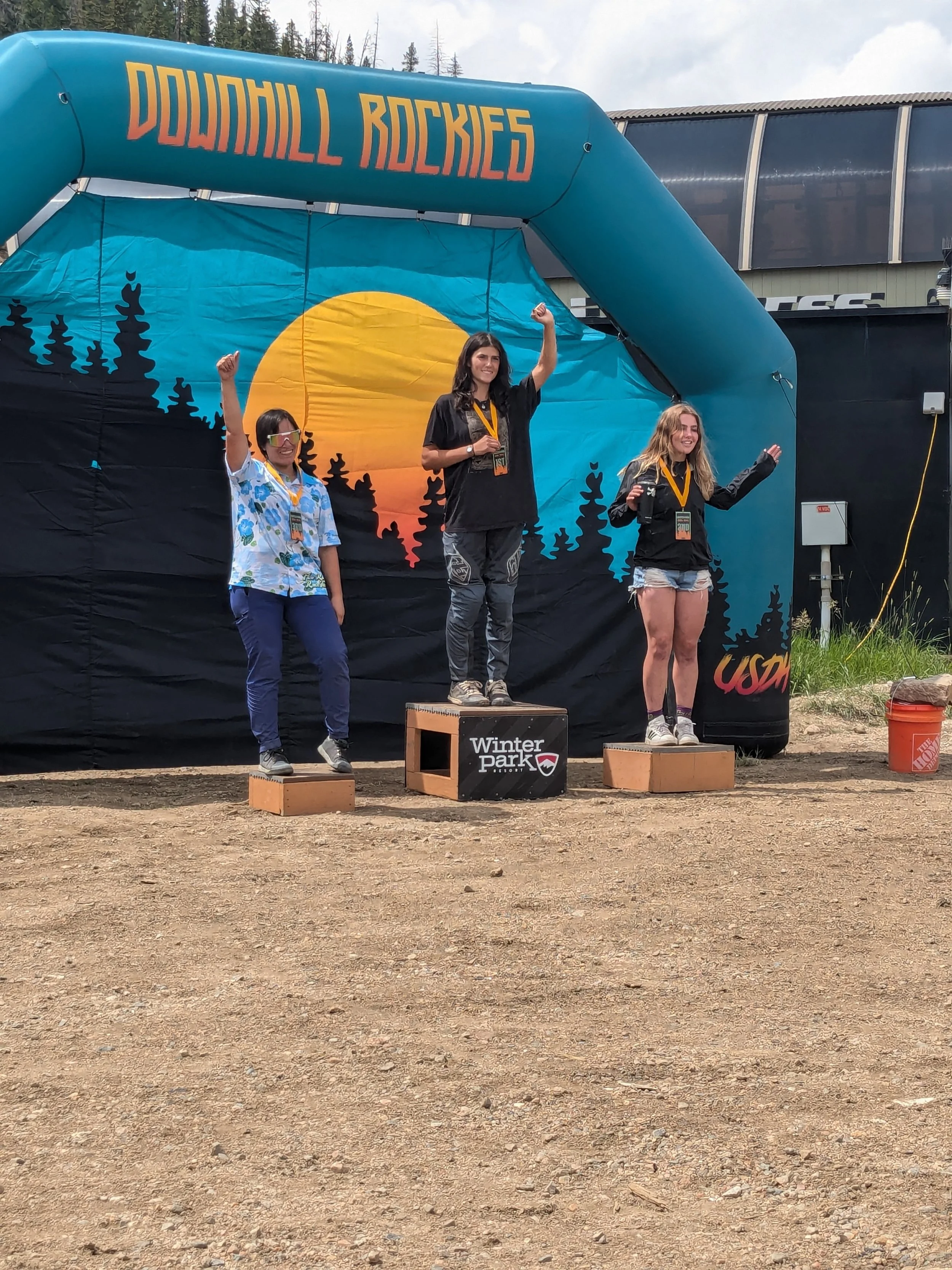 Three women standing on a winner's podium at a mountain biking event, with a backdrop of a mountain landscape and a large inflatable arch labeled 'Downhill Rockies.' The women are celebrating with raised fists and are wearing medals.