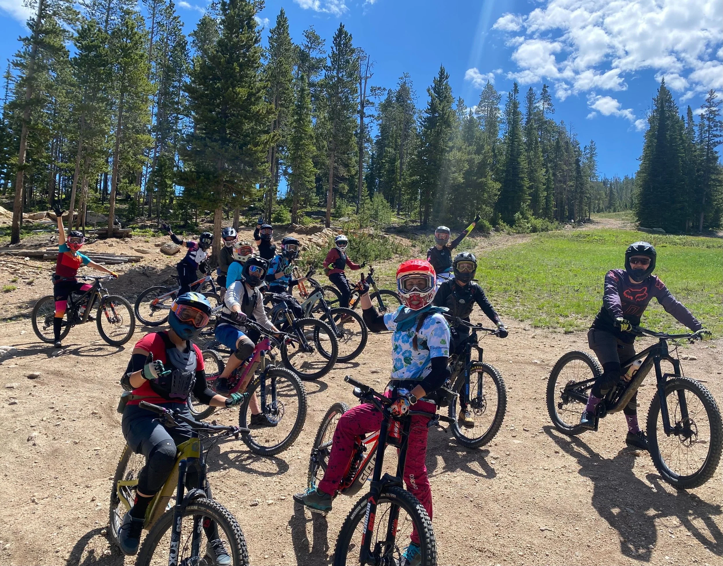 Group of children mountain biking on a dirt trail in a forested area on a sunny day, wearing helmets and protective gear.