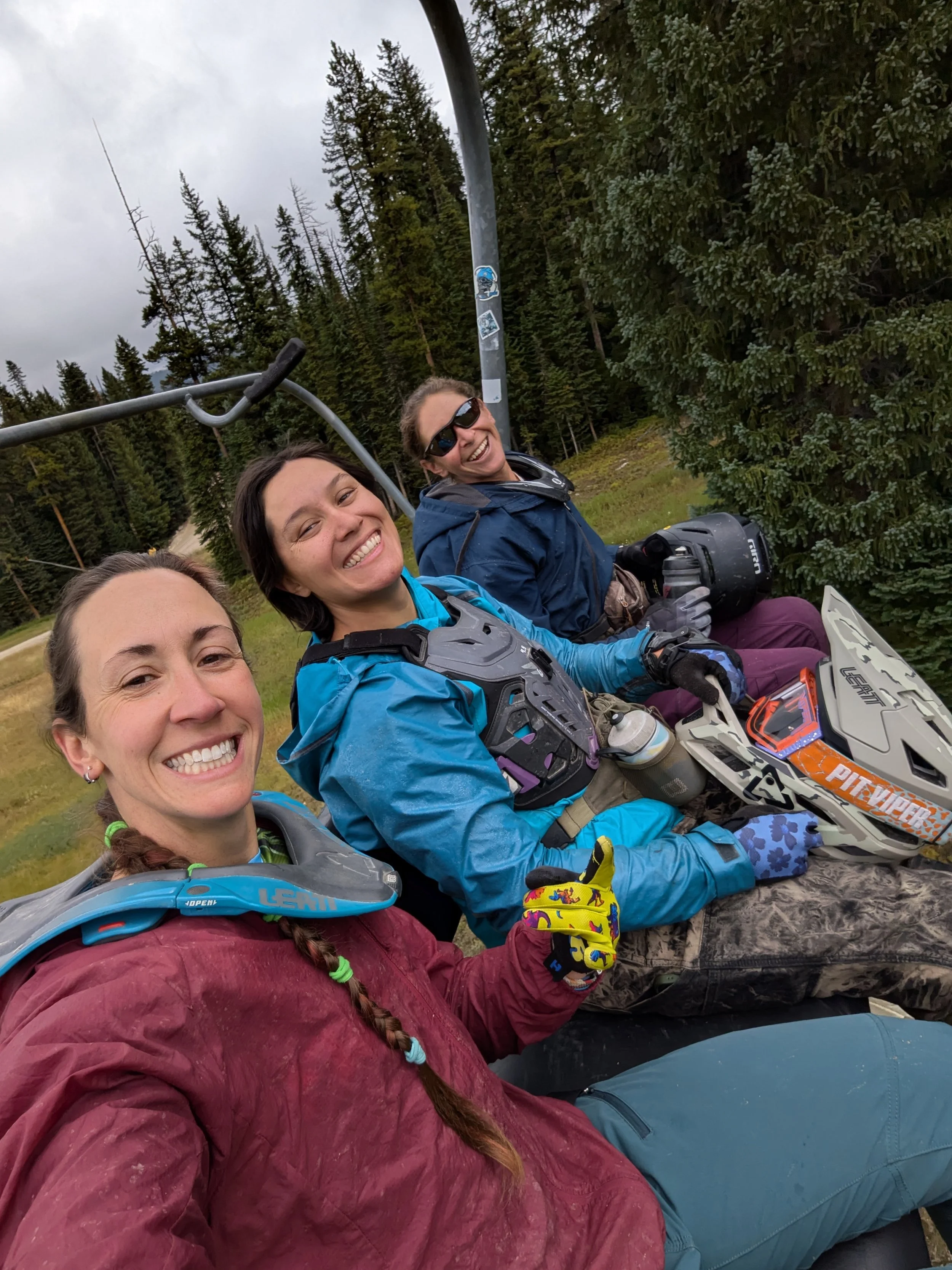 Three women sitting on a ski lift in a forested area, smiling and dressed in mountain biking gear, including helmets and gloves, with trees in the background.