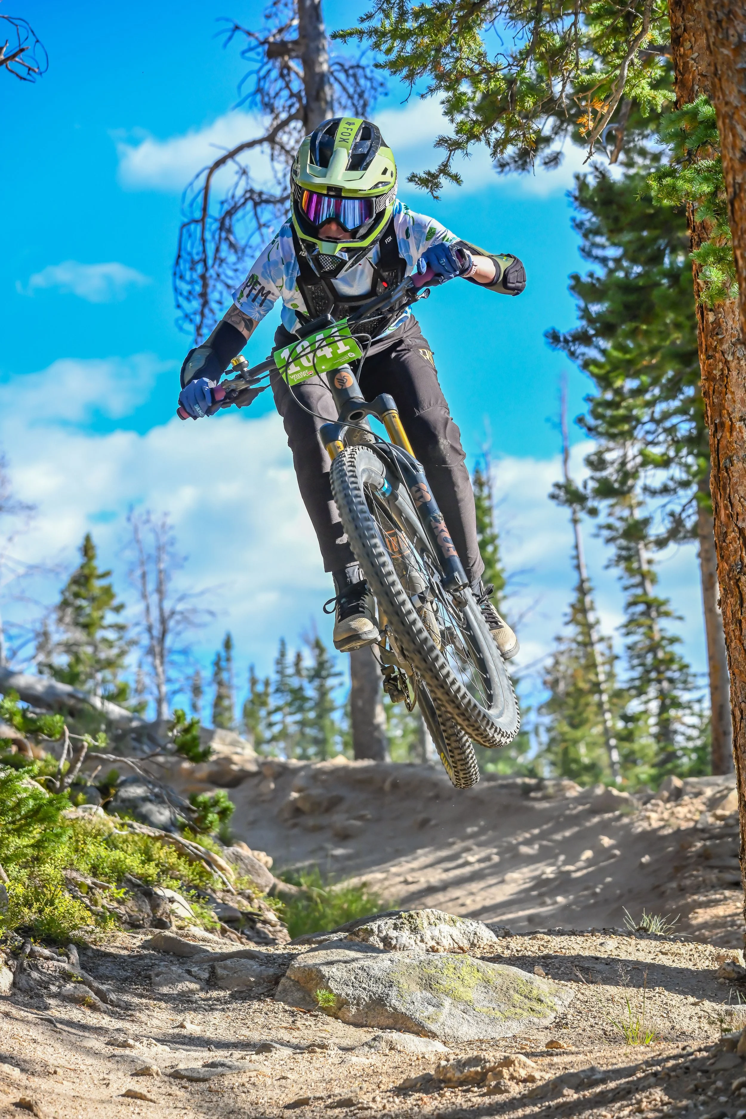 Mountain biker in mid-air jumping on a rocky trail through a forest with clear blue sky.