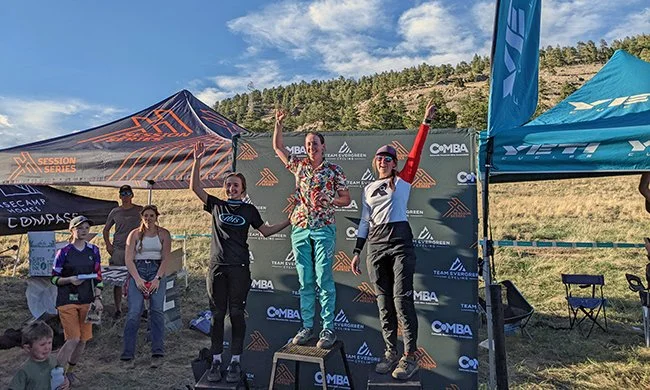 Three women on a winners' podium celebrating at an outdoor event, with tents and a scenic hillside in the background.