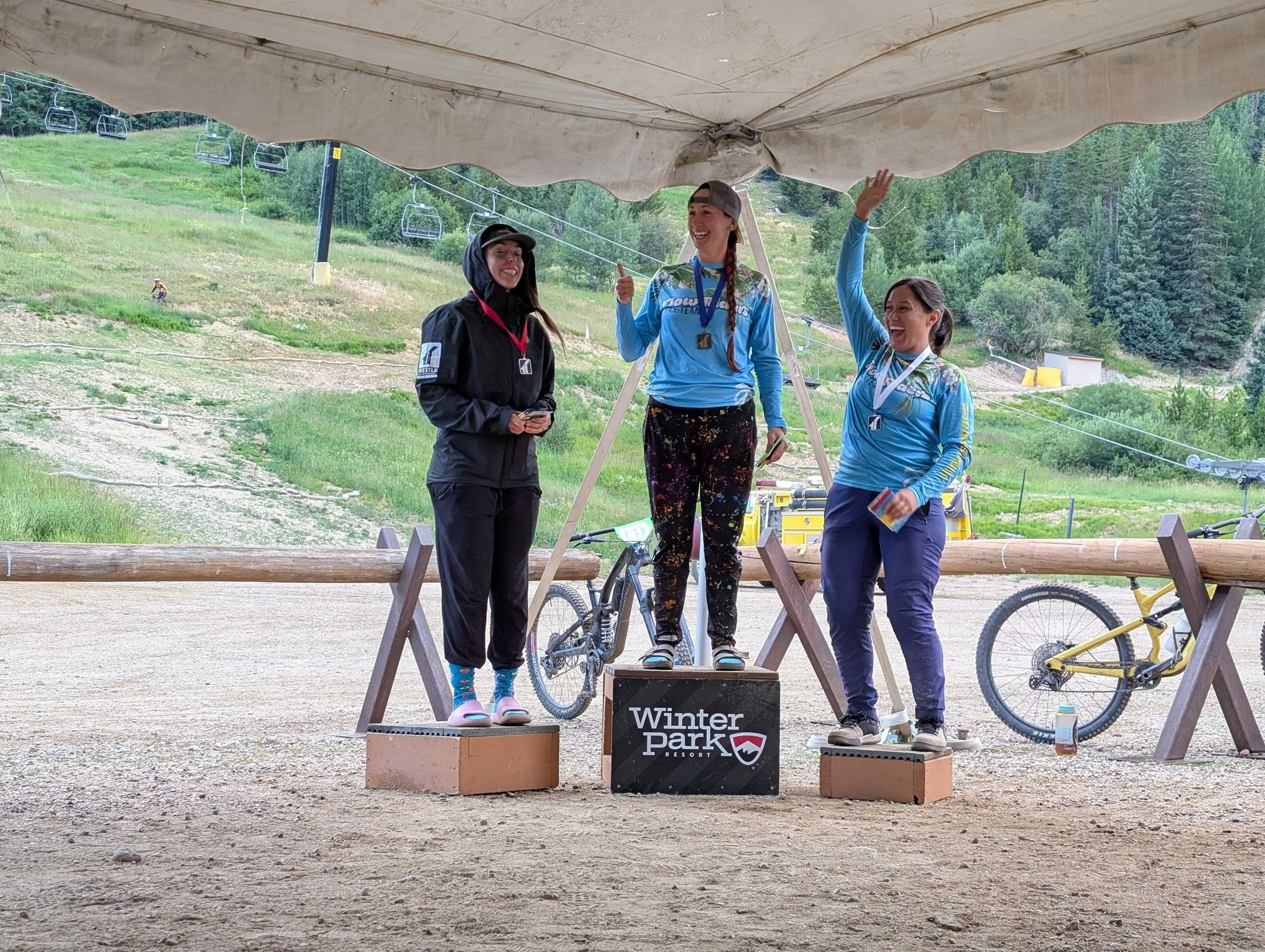 Three women standing on a winners' podium outdoors at Winter Park Resort, celebrating a competition with medals around their necks, surrounded by green mountains and ski lifts.