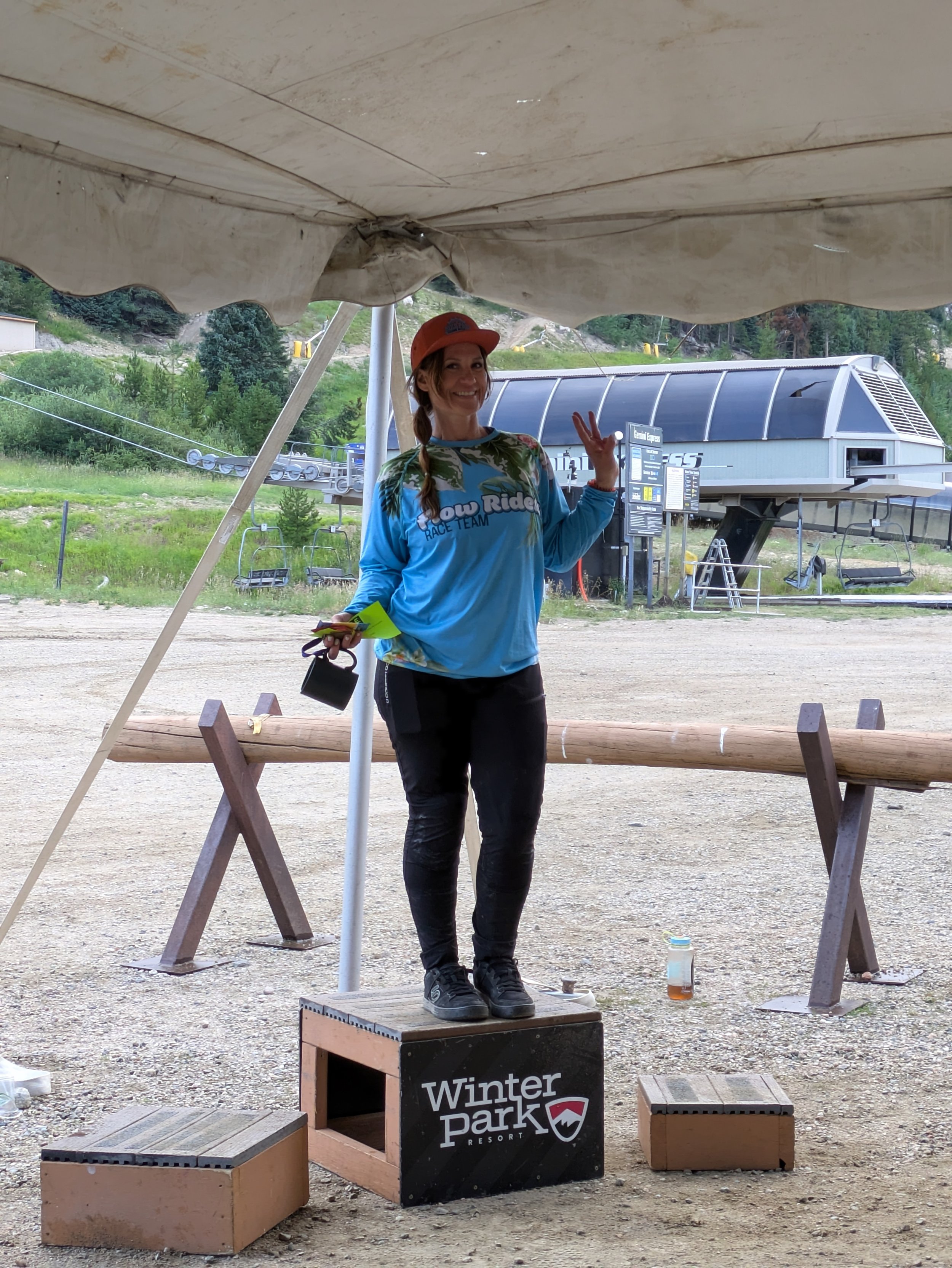 A woman standing on a winner's box at Winter Park Resort, wearing a blue race team jersey and an orange cap, smiling and waving.