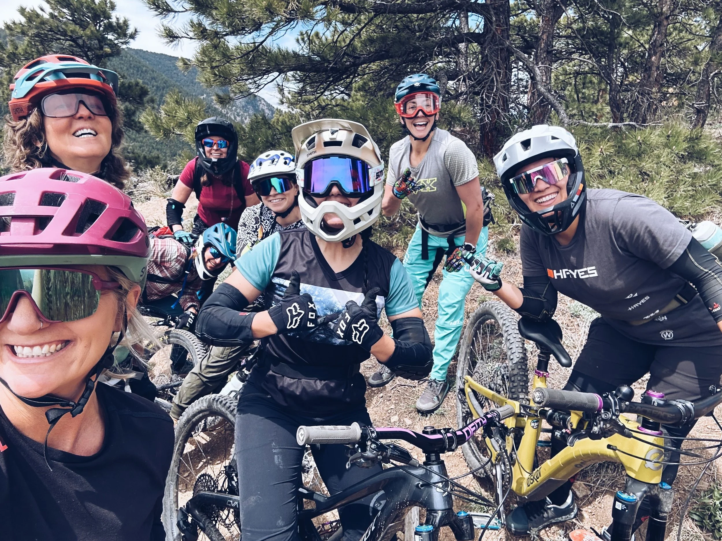 Group of women mountain biking on a trail surrounded by trees and mountains, all wearing helmets and protective gear.