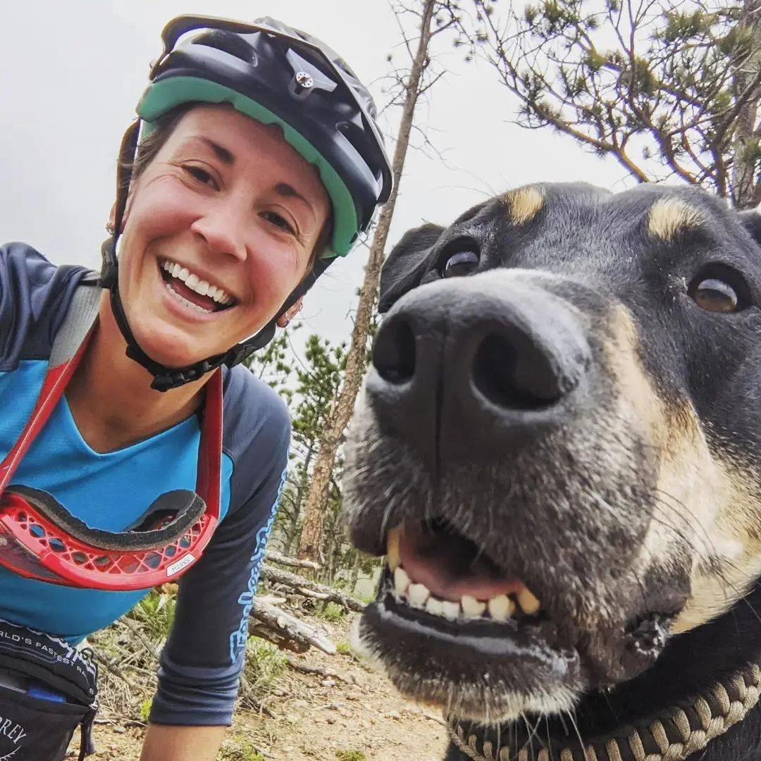 A woman with a bike helmet smiling for a selfie next to a large dog outdoors with trees and cloudy sky in the background.