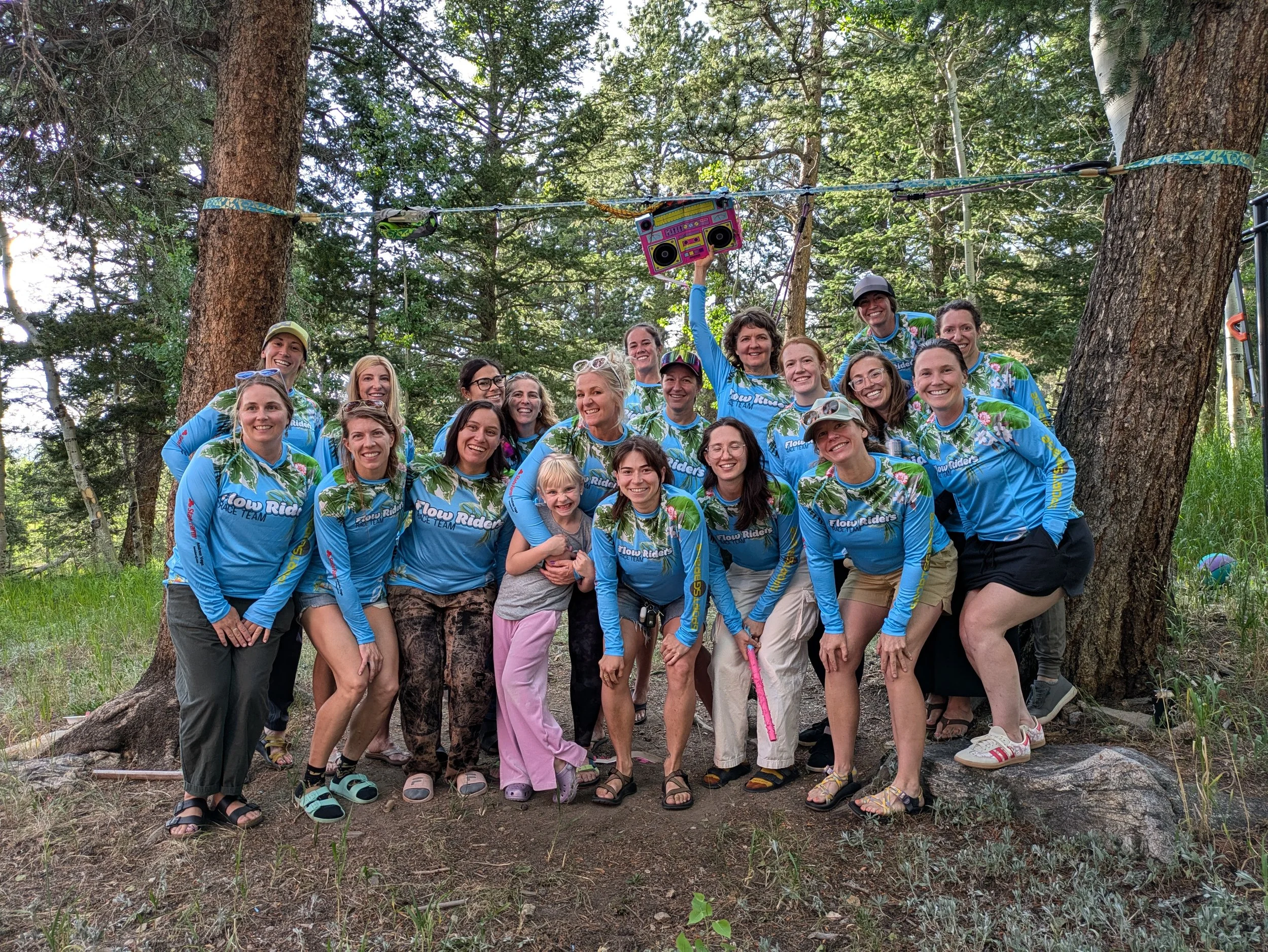 Group of people in matching blue flow rider shirts standing in a forest, smiling for a photo. Some are leaning on each other, one person is holding a colorful boombox above their head. They are surrounded by trees and natural scenery.