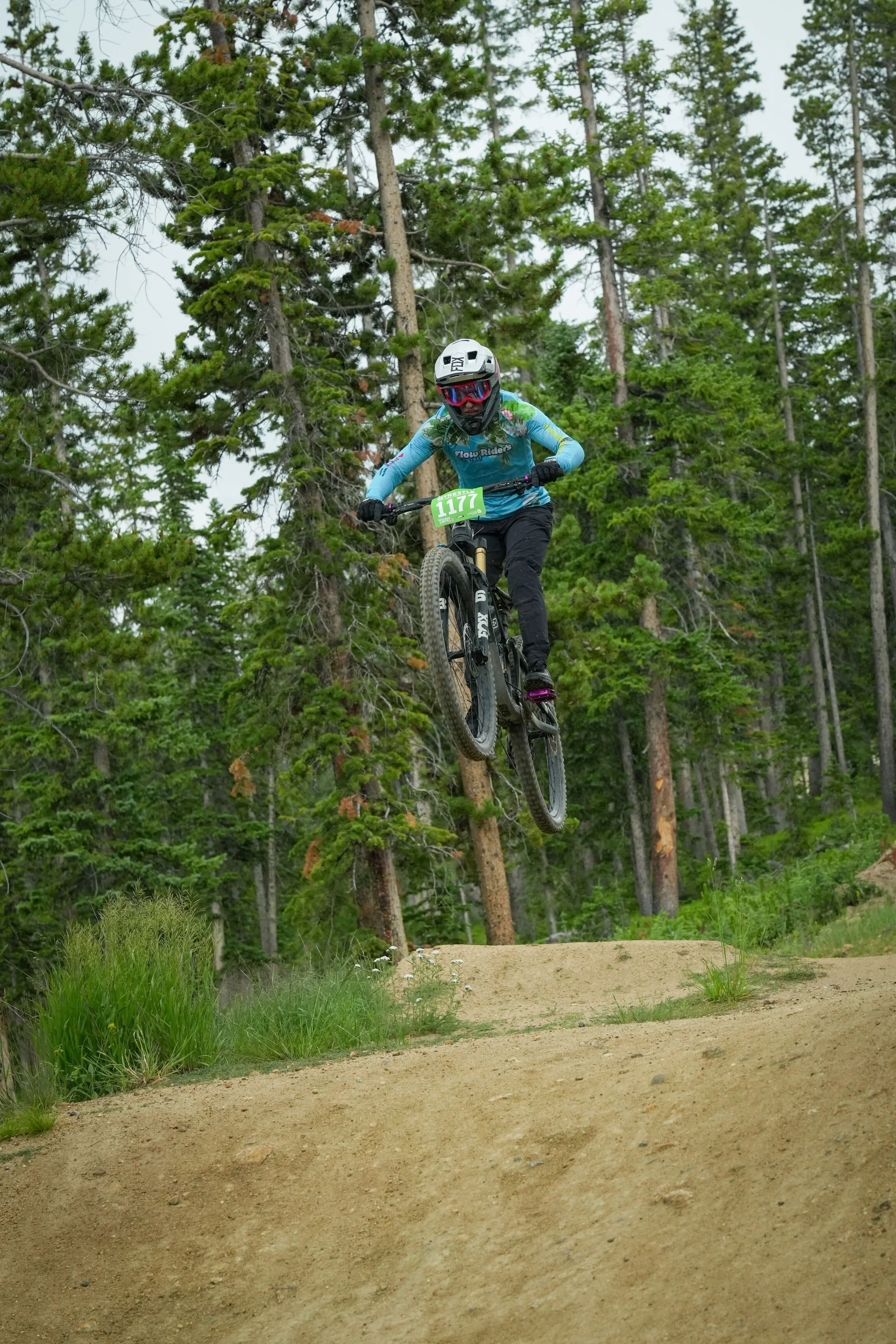Mountain biker mid-air going over a jump on a forest trail.