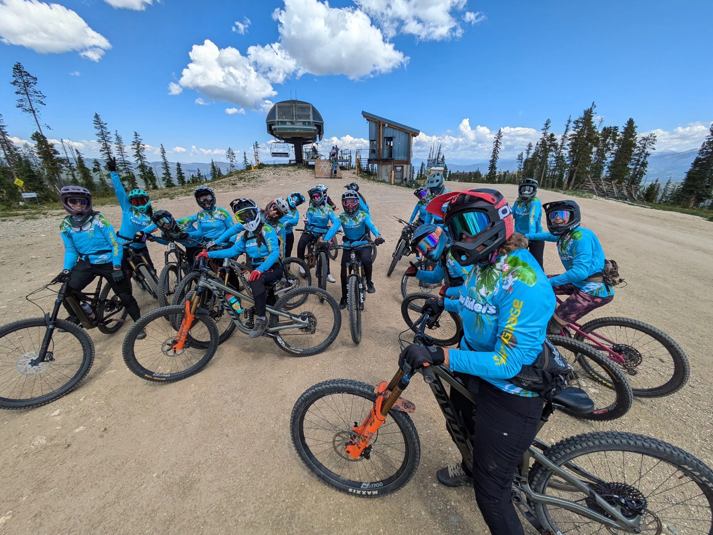 Group of cyclists wearing blue jerseys and helmets, posing with their mountain bikes on a dirt trail at a mountain resort, with ski lift structures and trees in the background under a clear sky with some clouds.