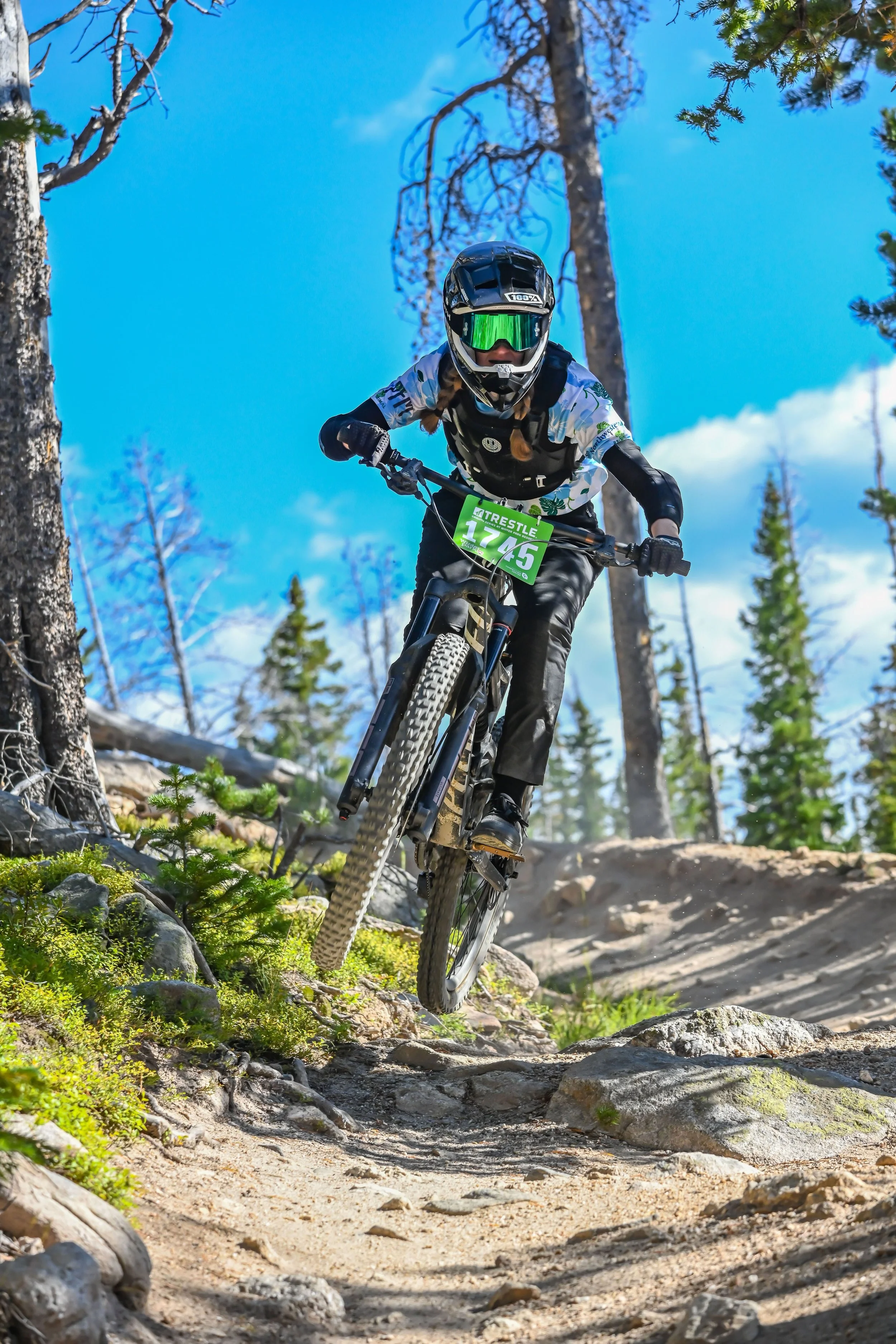 Mountain biker wearing a helmet and goggles riding downhill on rocky trail in forest with pine trees and blue sky.