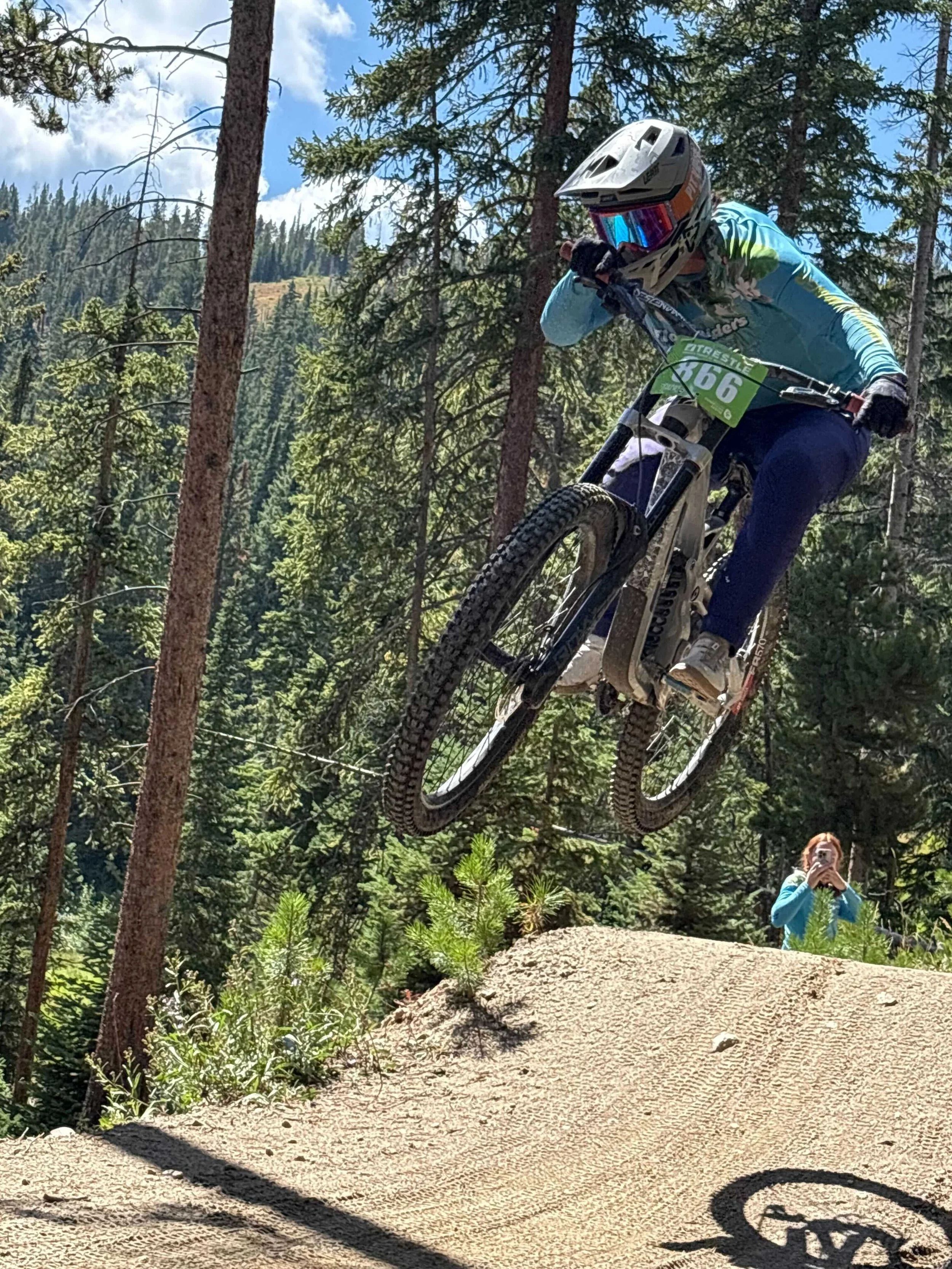 A mountain biker wearing a helmet and goggles jumping off a dirt ledge on a forest trail, with trees and a person taking a photo in the background.