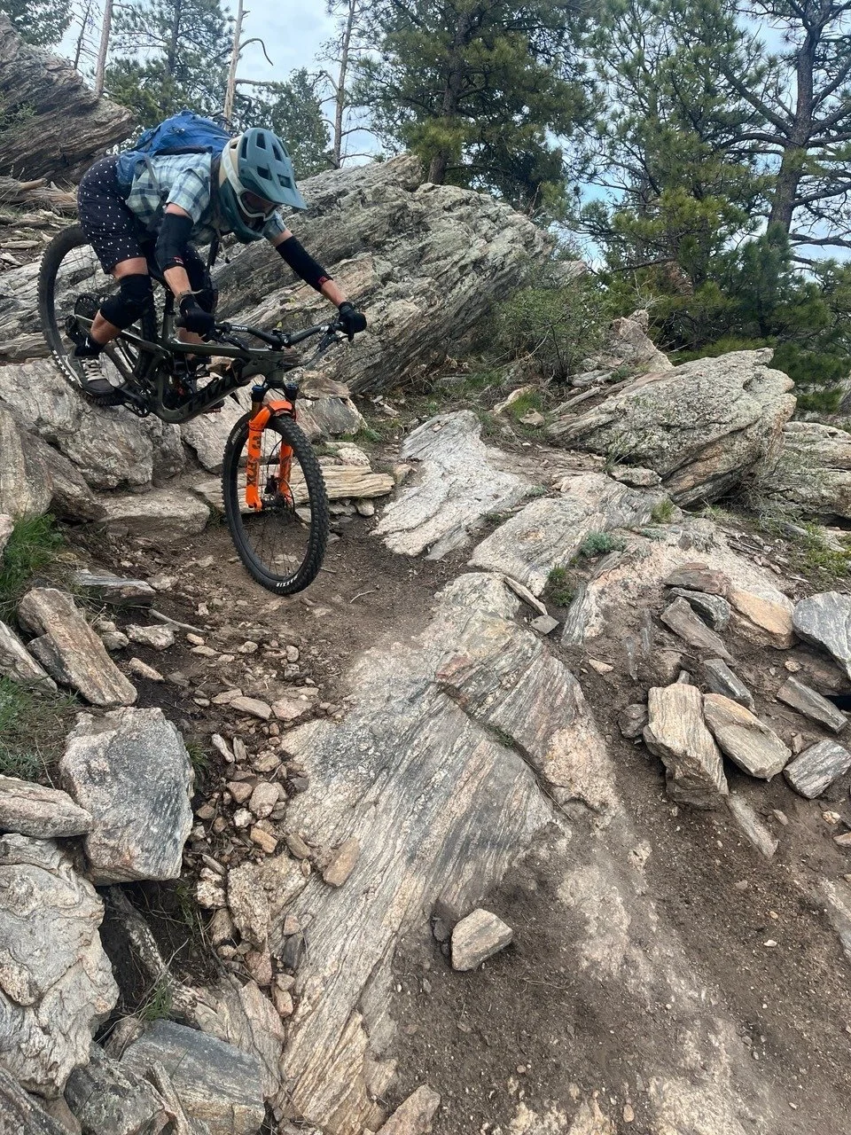 A mountain biker wearing a helmet, gloves, and sunglasses navigating a rocky downhill trail surrounded by trees and large rocks.